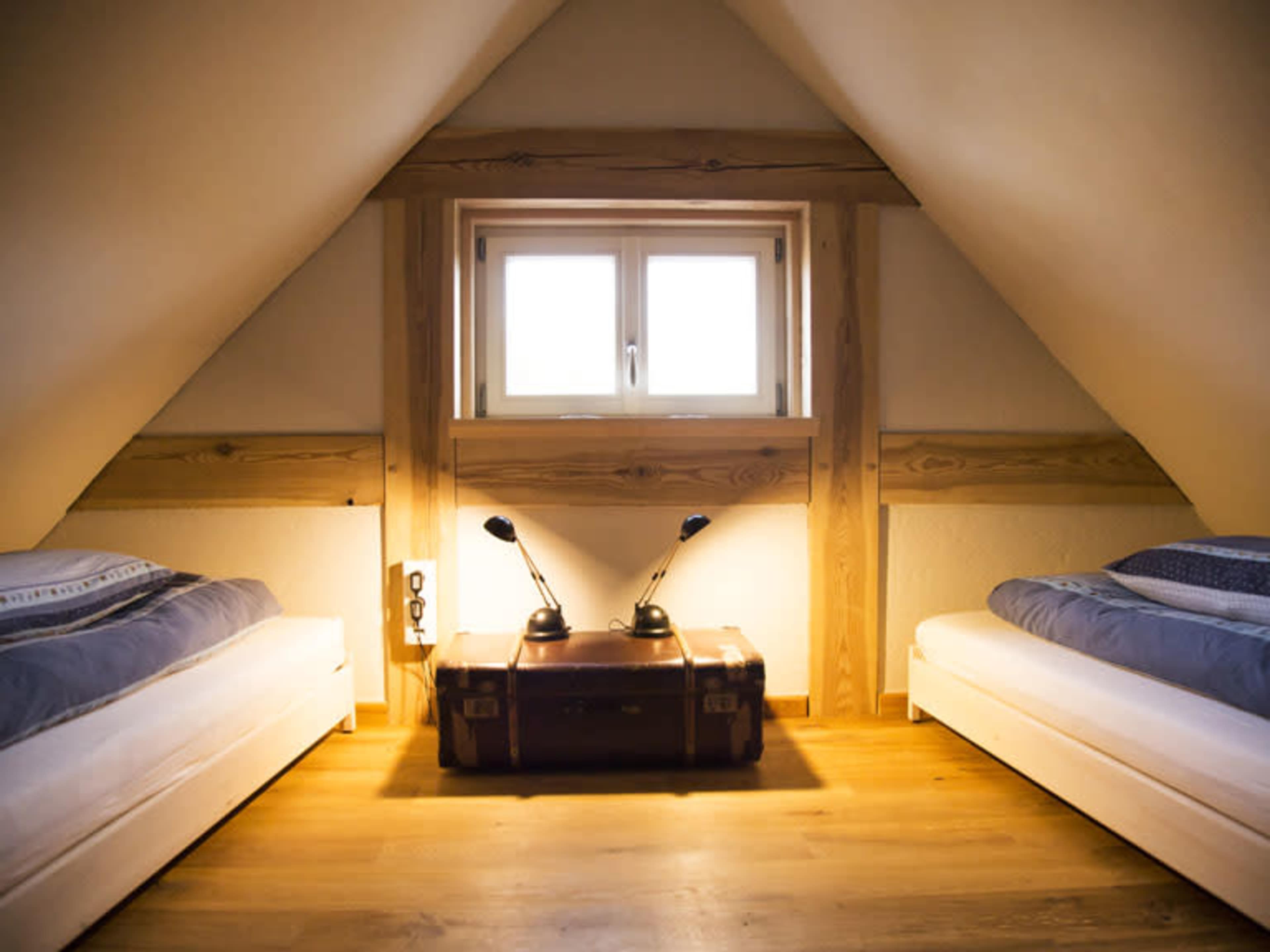 A cozy attic room features two single beds placed on either side of a wooden trunk, with a window above and two desk lamps positioned on the trunk.