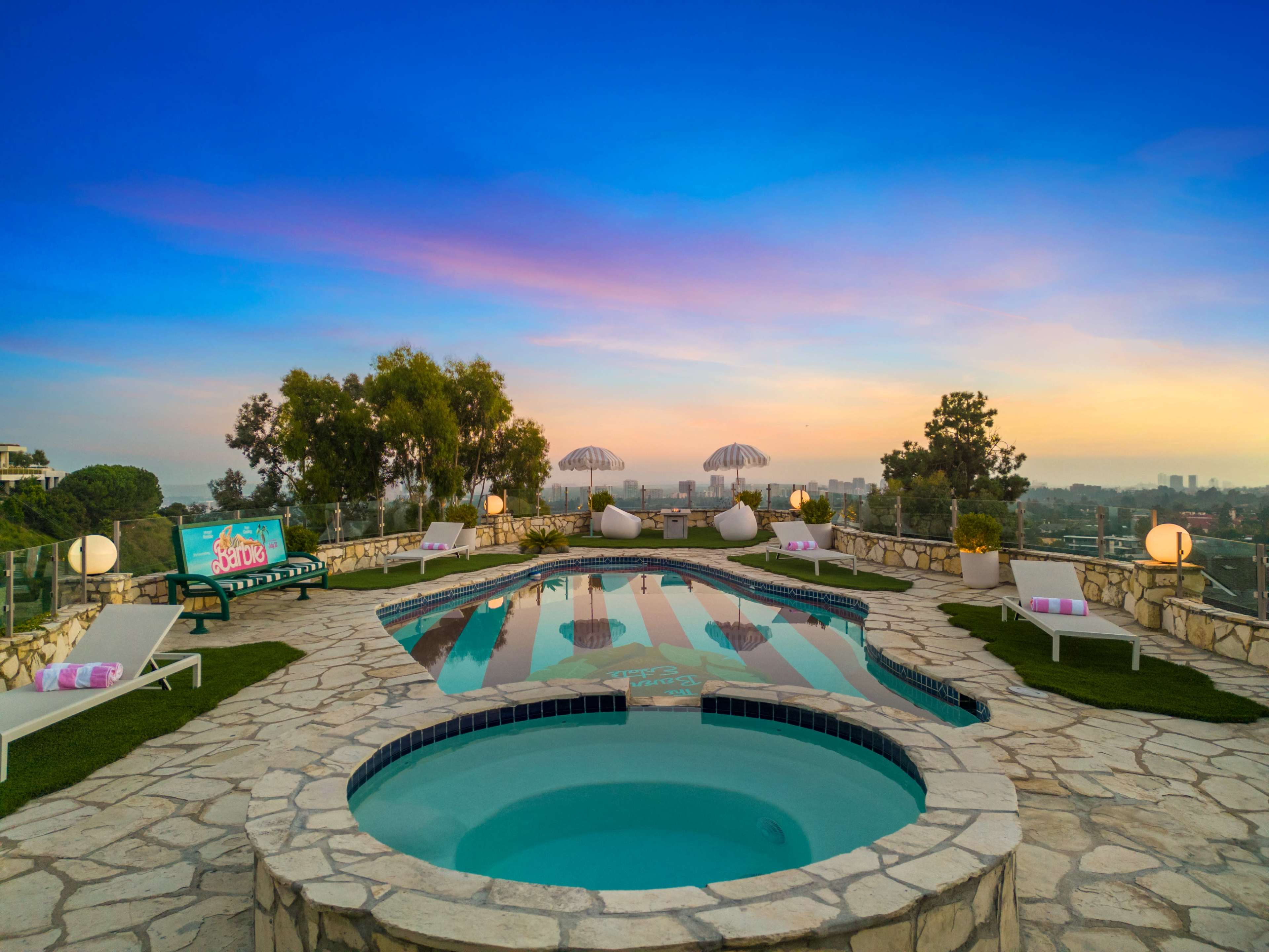 A hillside pool area featuring a large swimming pool, a smaller hot tub, and lounge chairs under parasols, set against a colorful sunset sky.