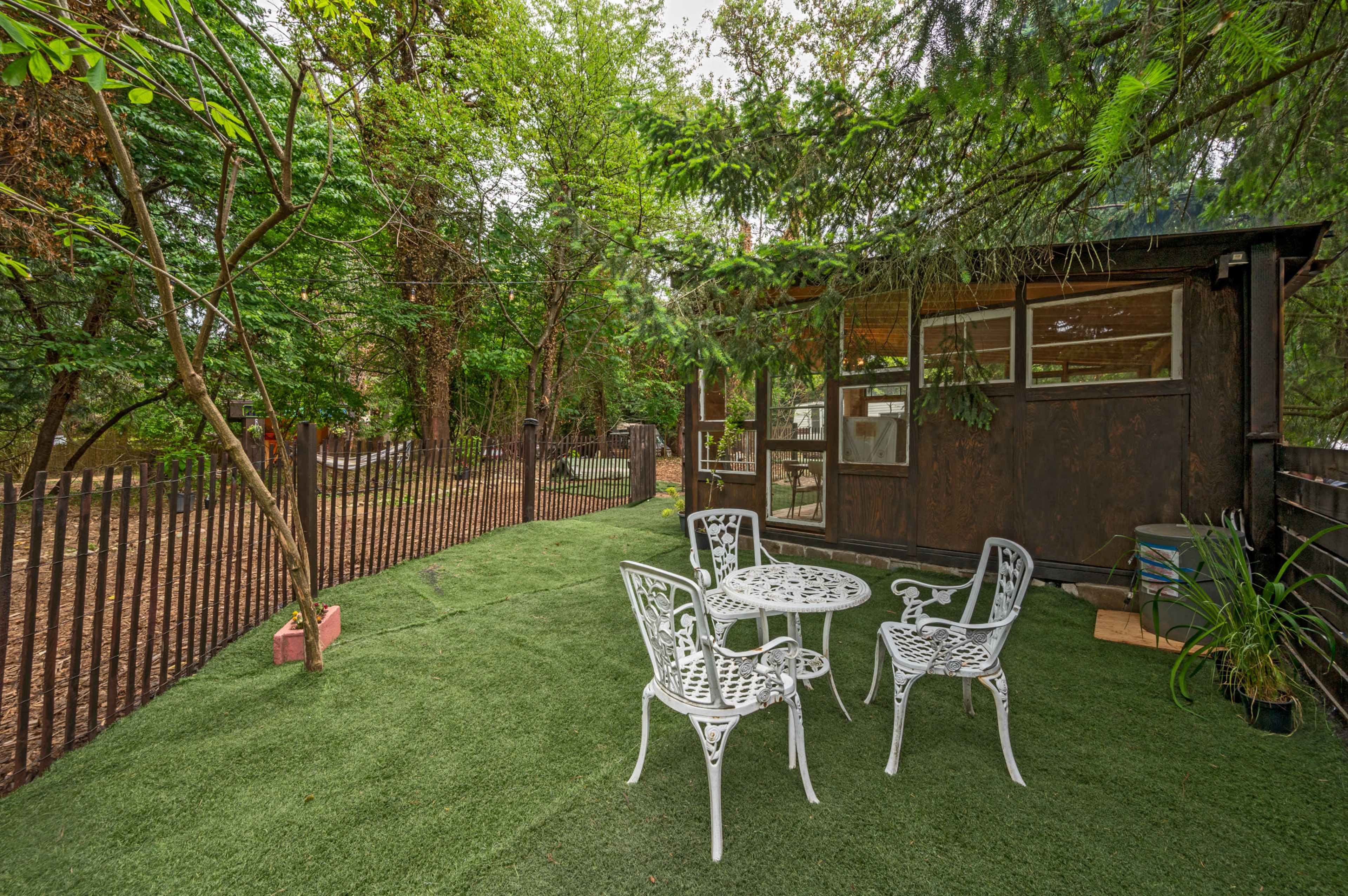 A small outdoor seating area with white metal furniture is set on artificial grass near a wooden cabin surrounded by trees.