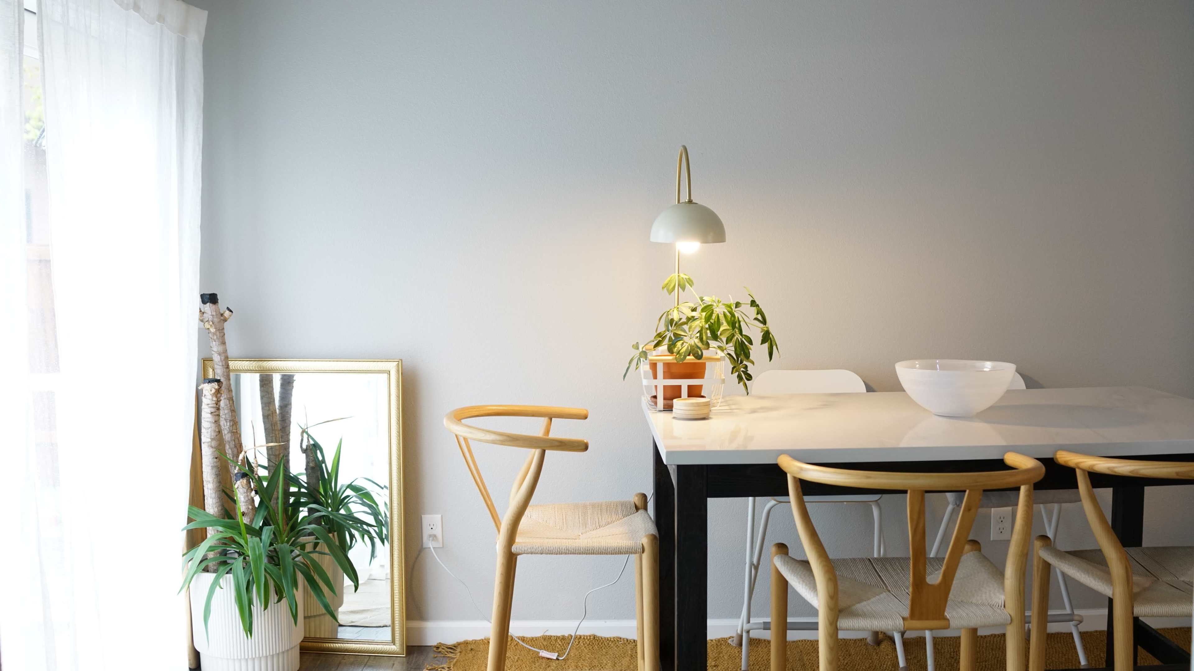 A dining area features a white table, wooden chairs, a potted plant, and a mirror against a light gray wall.