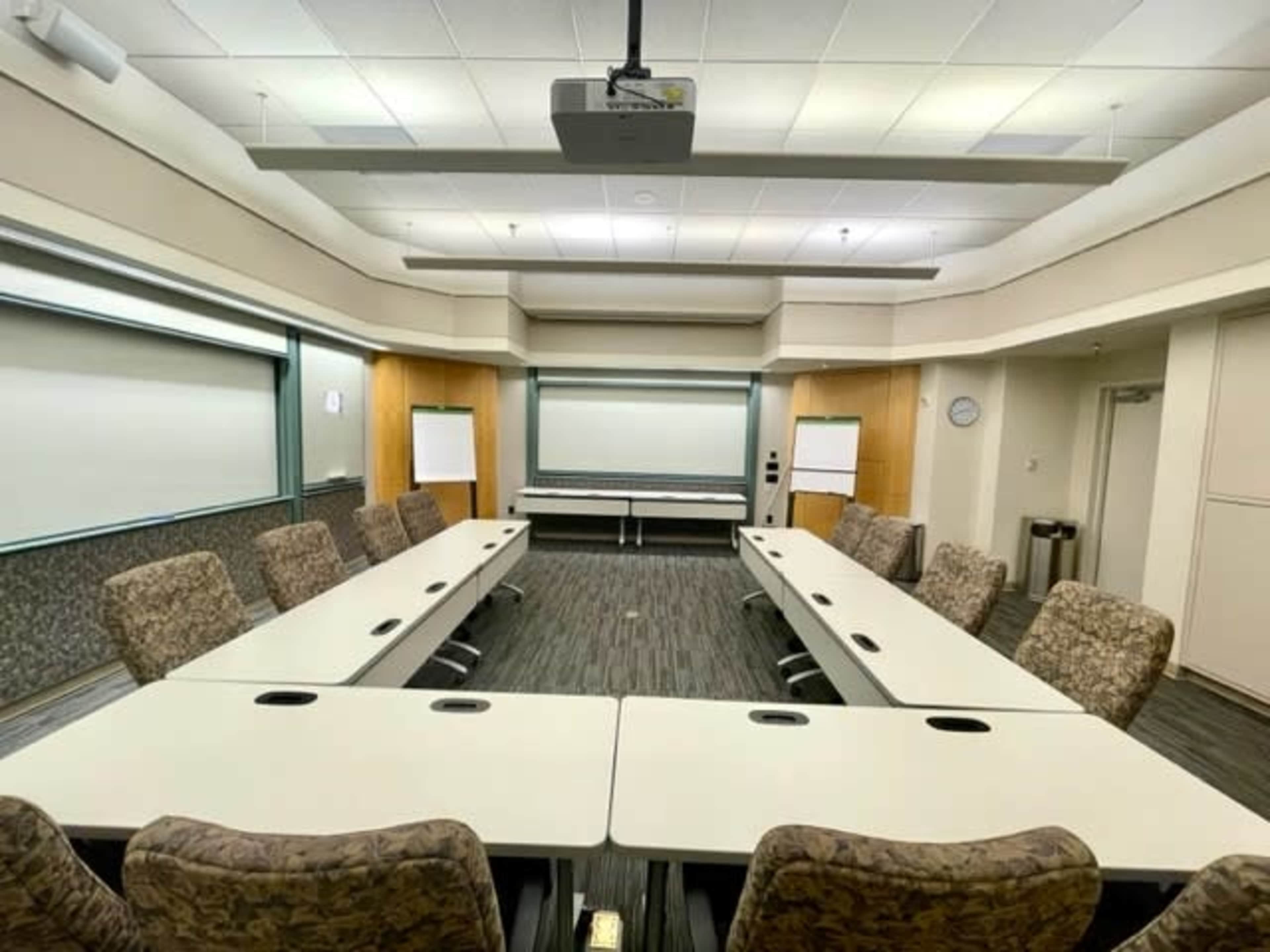 A conference room with a large oval table surrounded by upholstered chairs and a projector mounted on the ceiling.