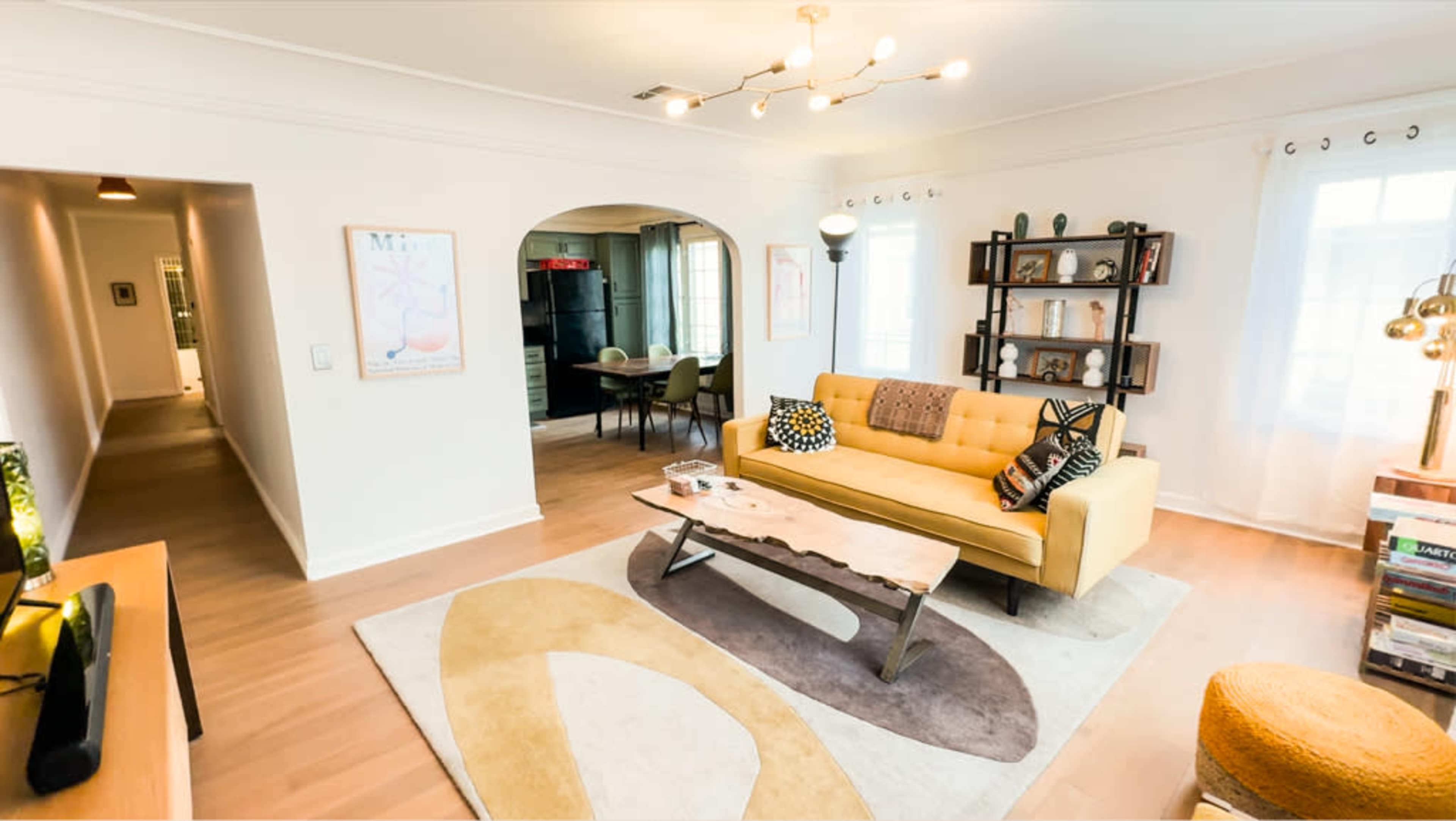 The image shows a bright living room with a yellow sofa, a wooden coffee table, and a bookshelf, leading to an open hallway and dining area.