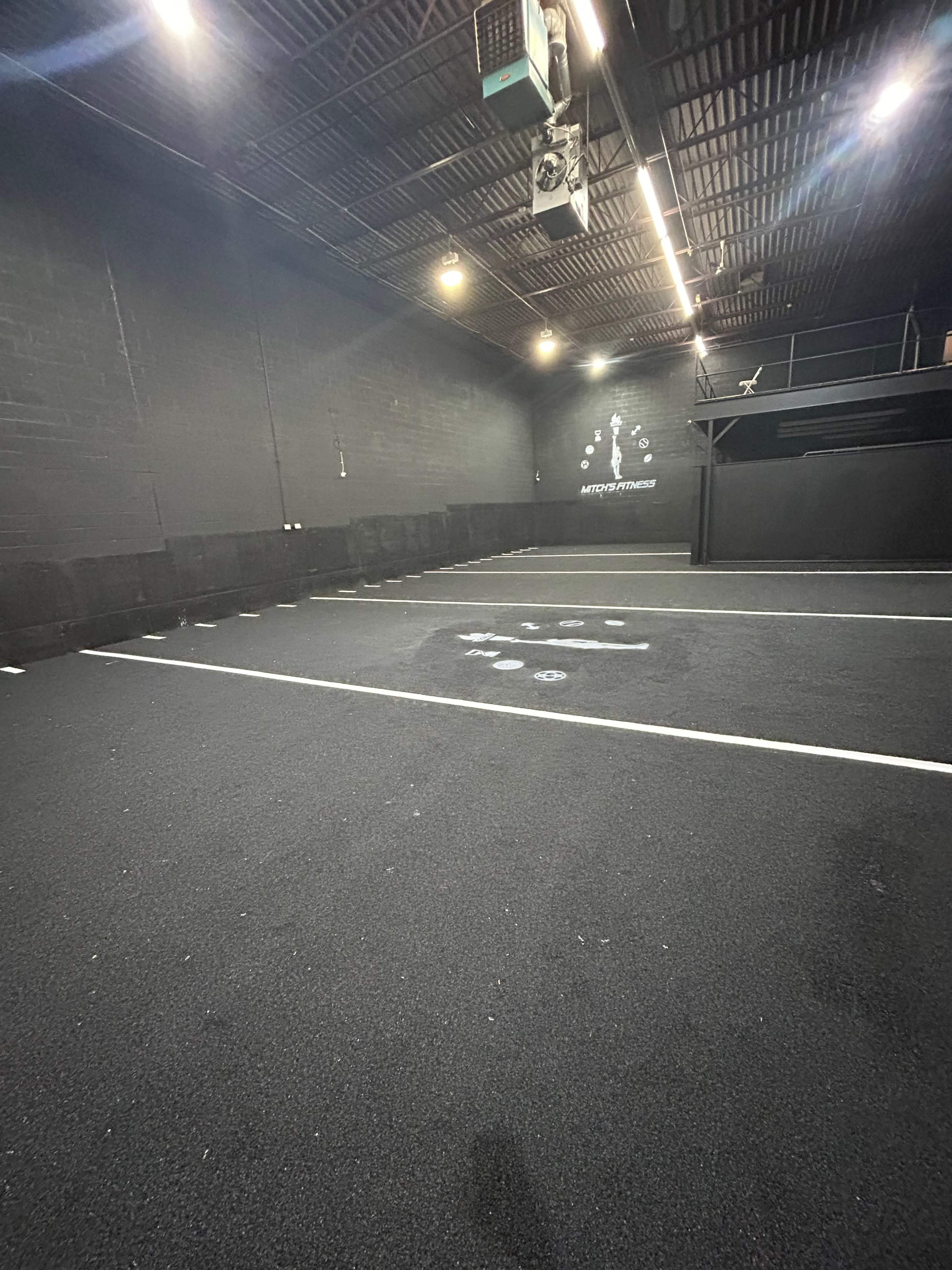 A spacious black rubber-floored gym with marked lines and a few exercise stations in a dimly lit environment.