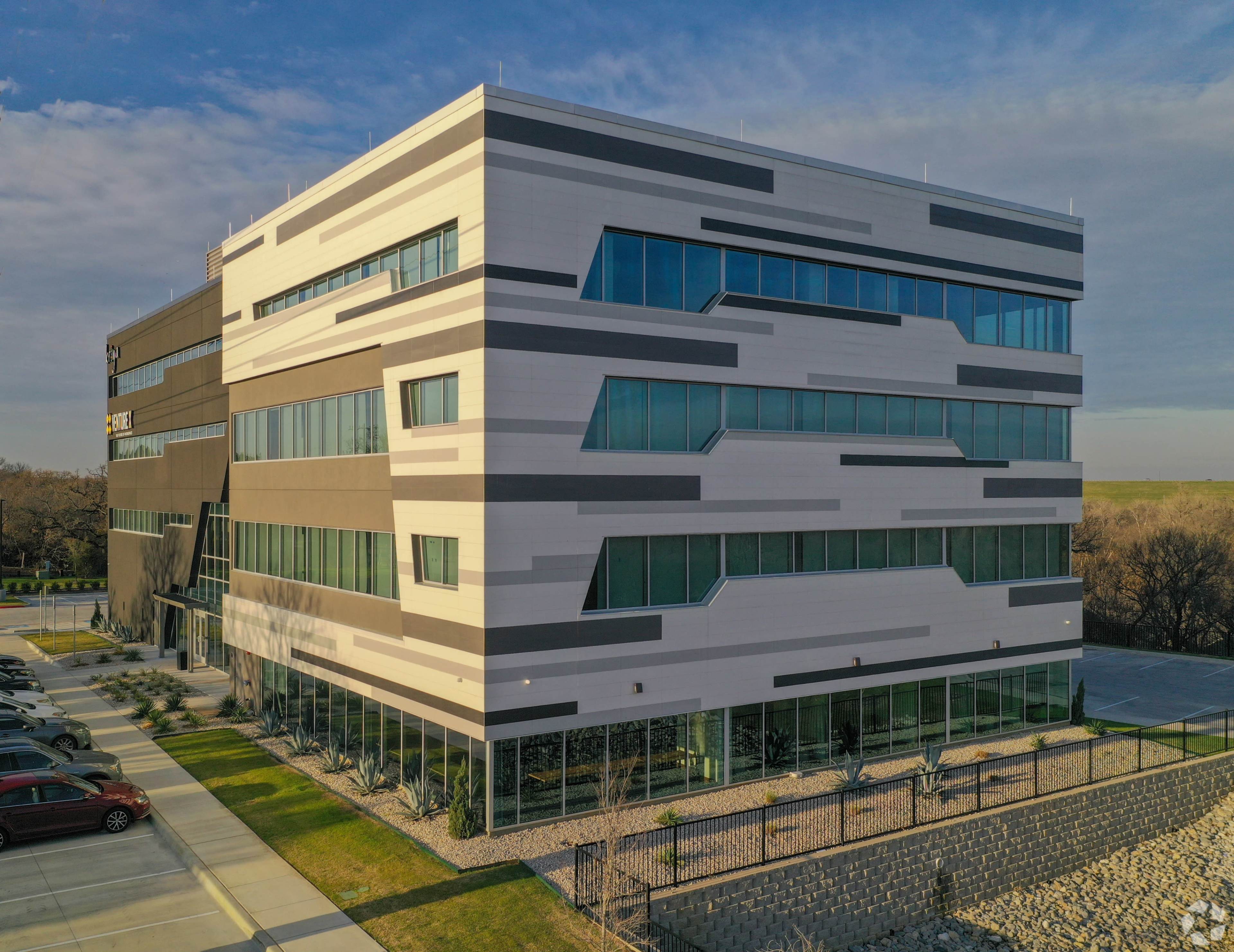A modern, multi-story building with angular architectural features and large windows, set in a landscaped area with parked cars nearby.
