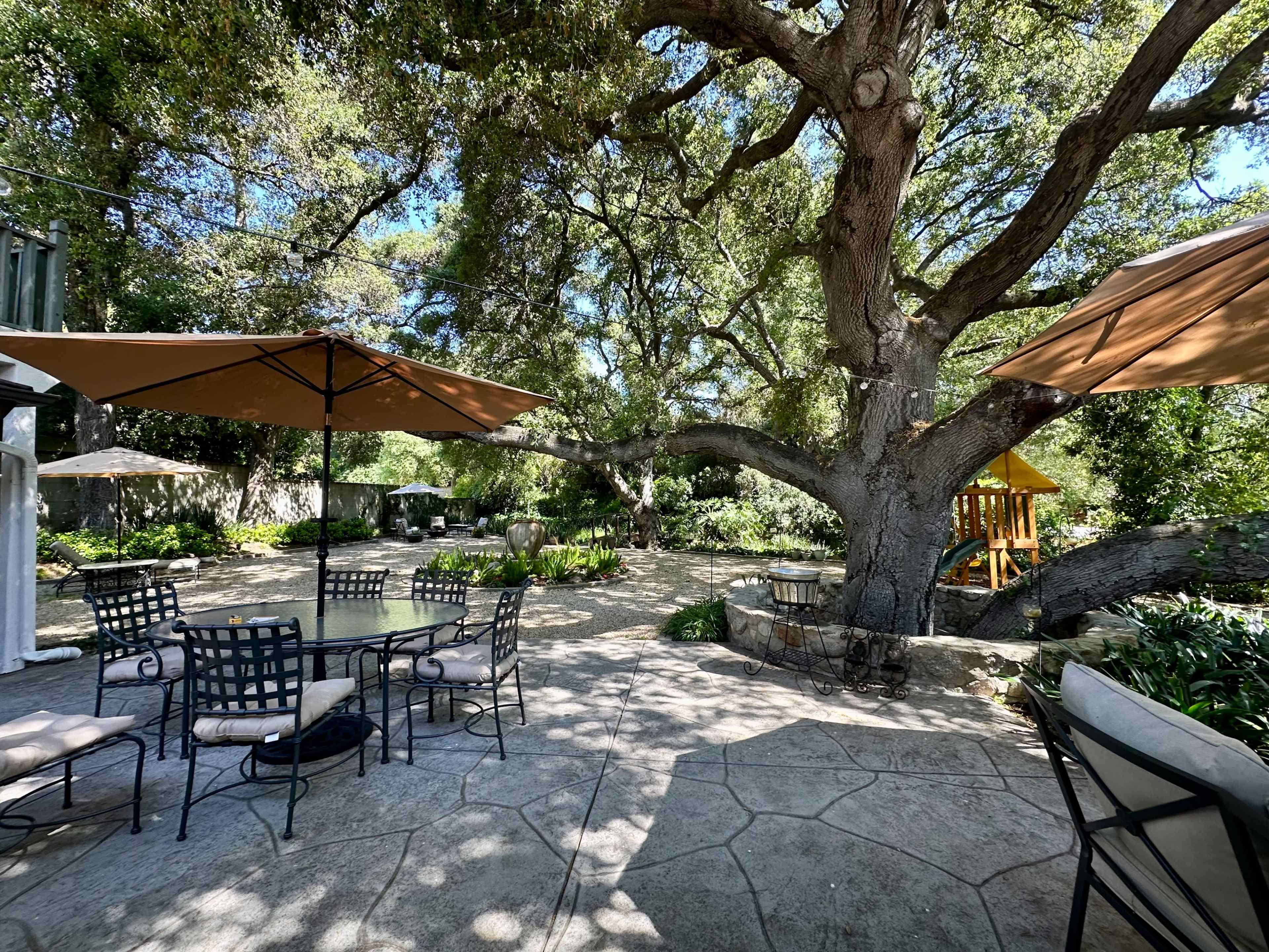 The image depicts a shaded outdoor patio area with a large tree, patio furniture, and a children's play structure in a landscaped garden.