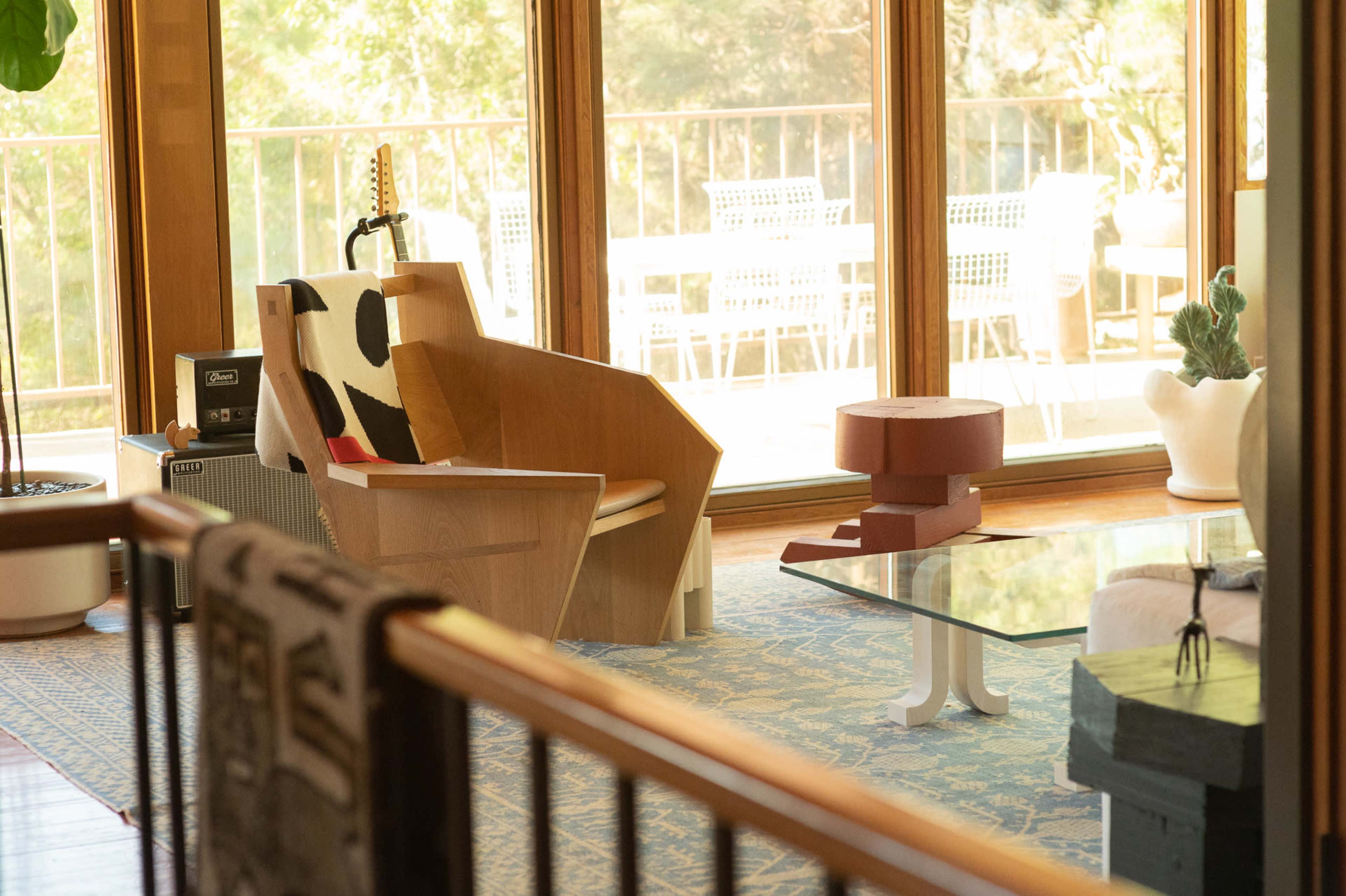 A modern living space featuring a wooden chair, a glass coffee table, and large windows overlooking a patio with outdoor seating.