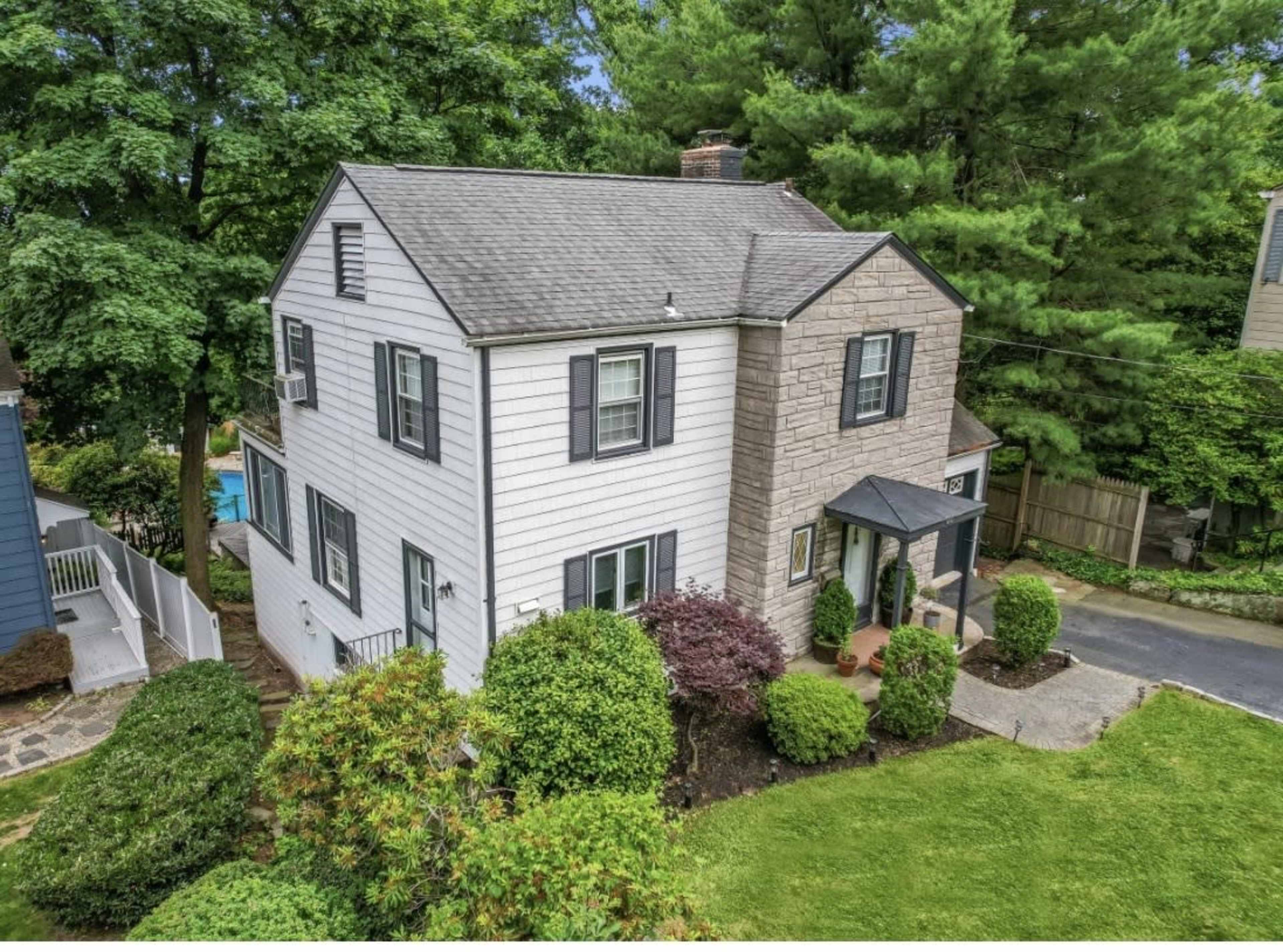 The image shows a two-story house with a mix of white siding and stonework, surrounded by well-maintained greenery and a clear walkway leading to the entrance.