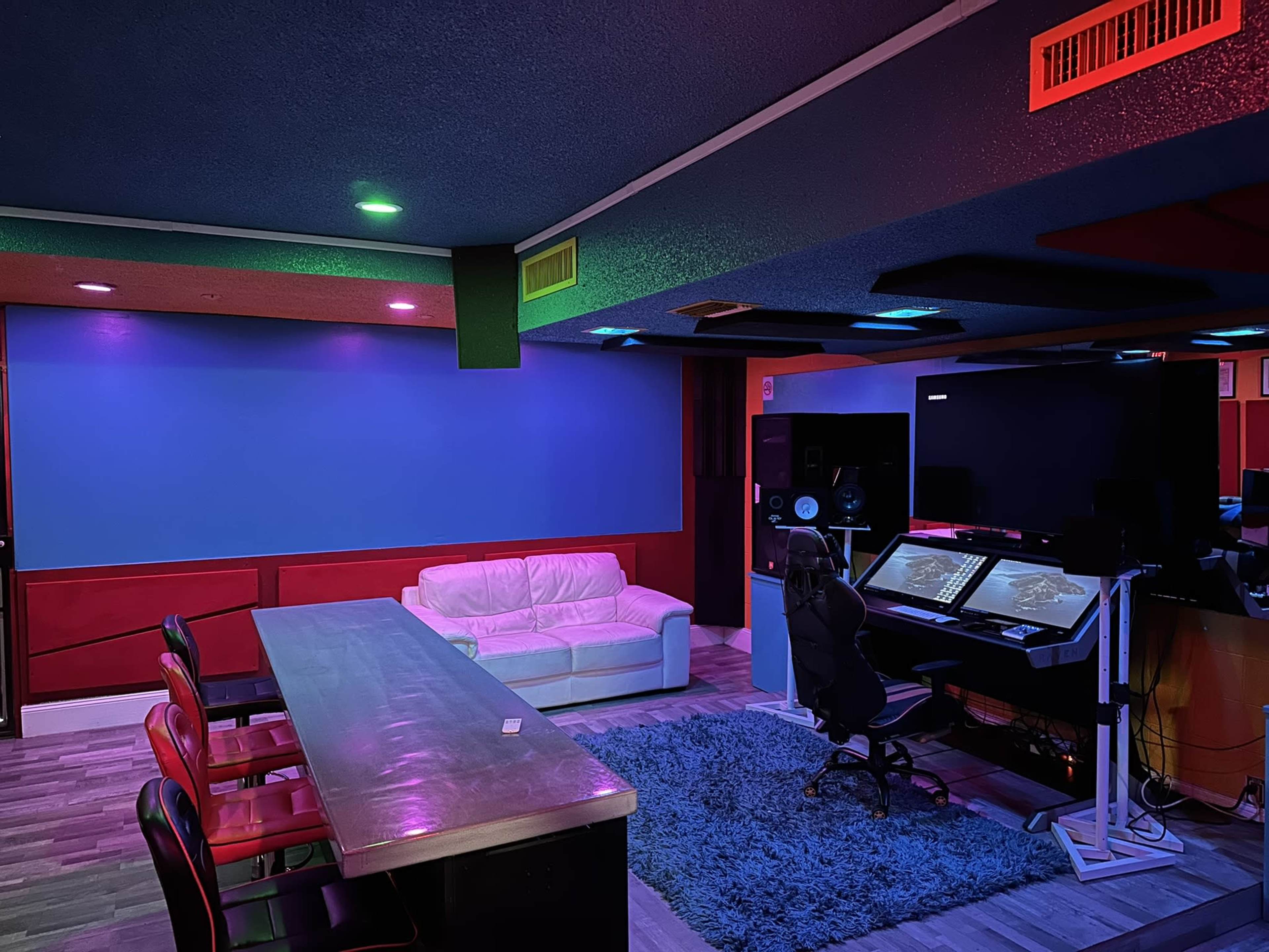 The scene features a modern recording studio with a white couch, a long table, and a desk equipped with music production equipment, all illuminated by colorful lights.