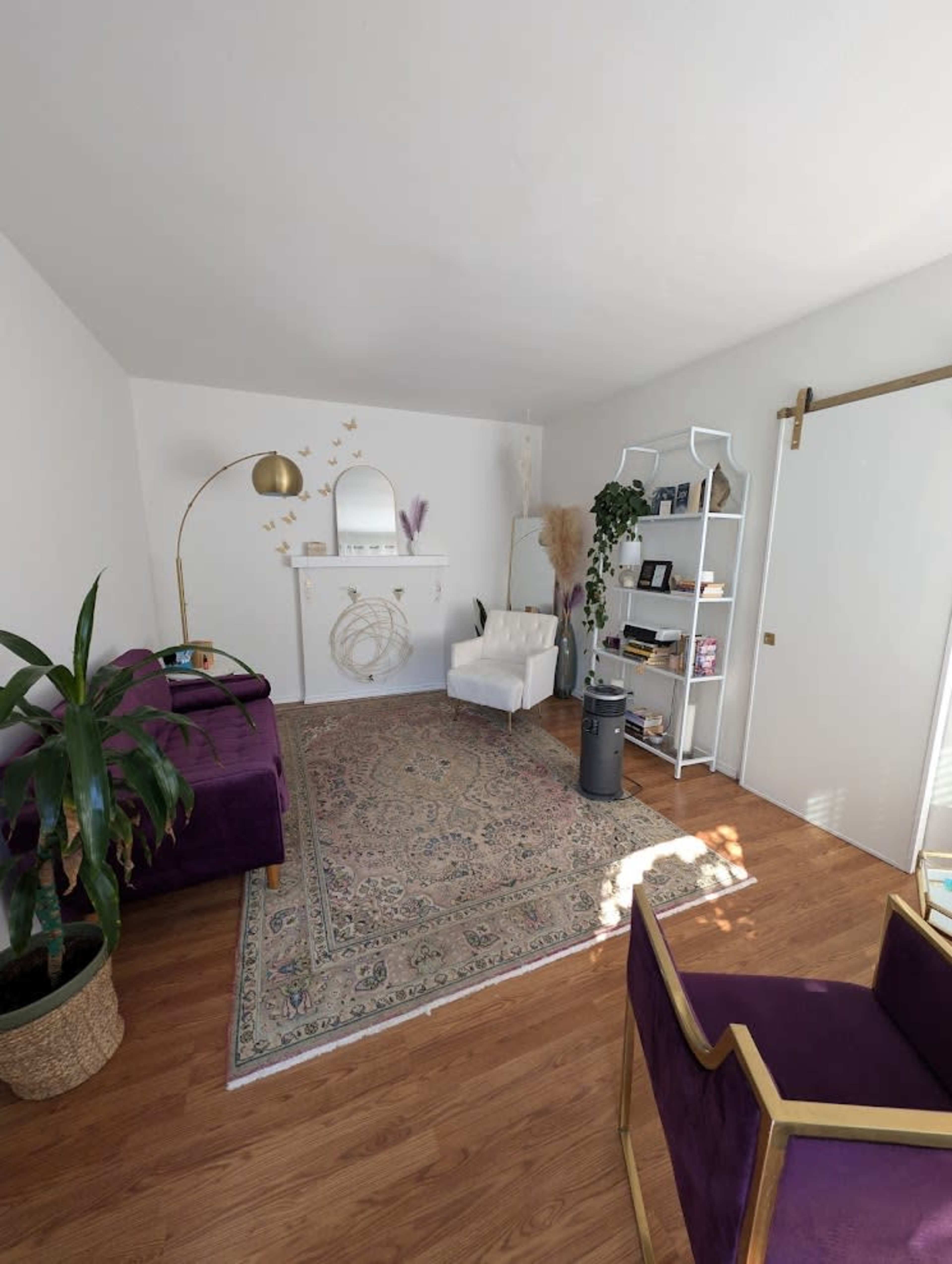 The image depicts a modern interior room featuring a purple couch, white armchair, decorative shelf, and a fireplace surrounded by plants and a patterned rug.