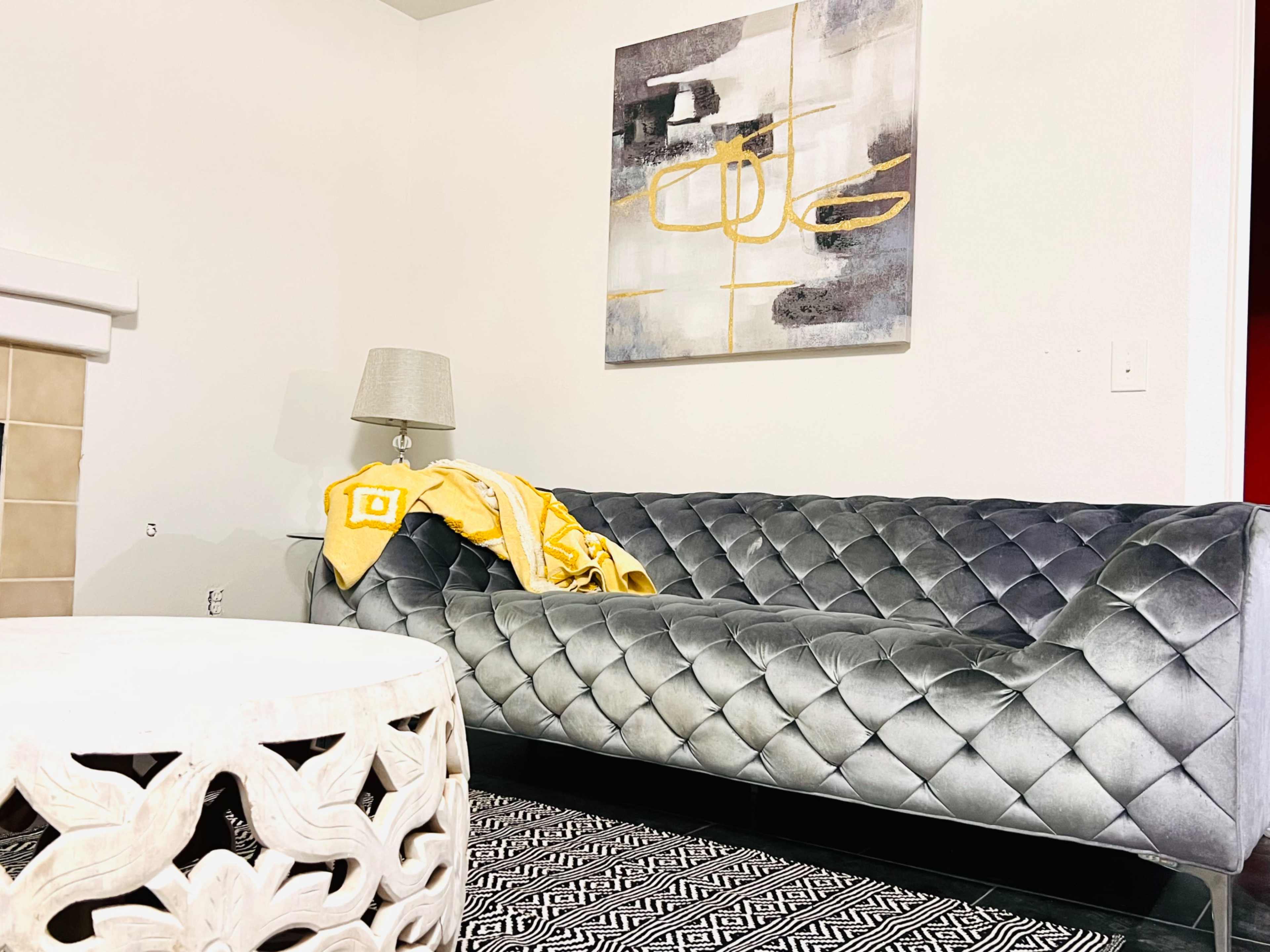 A tufted gray sofa is positioned next to a white lamp and a small decorative table, with a yellow blanket draped over the couch and a modern artwork hanging on the wall.