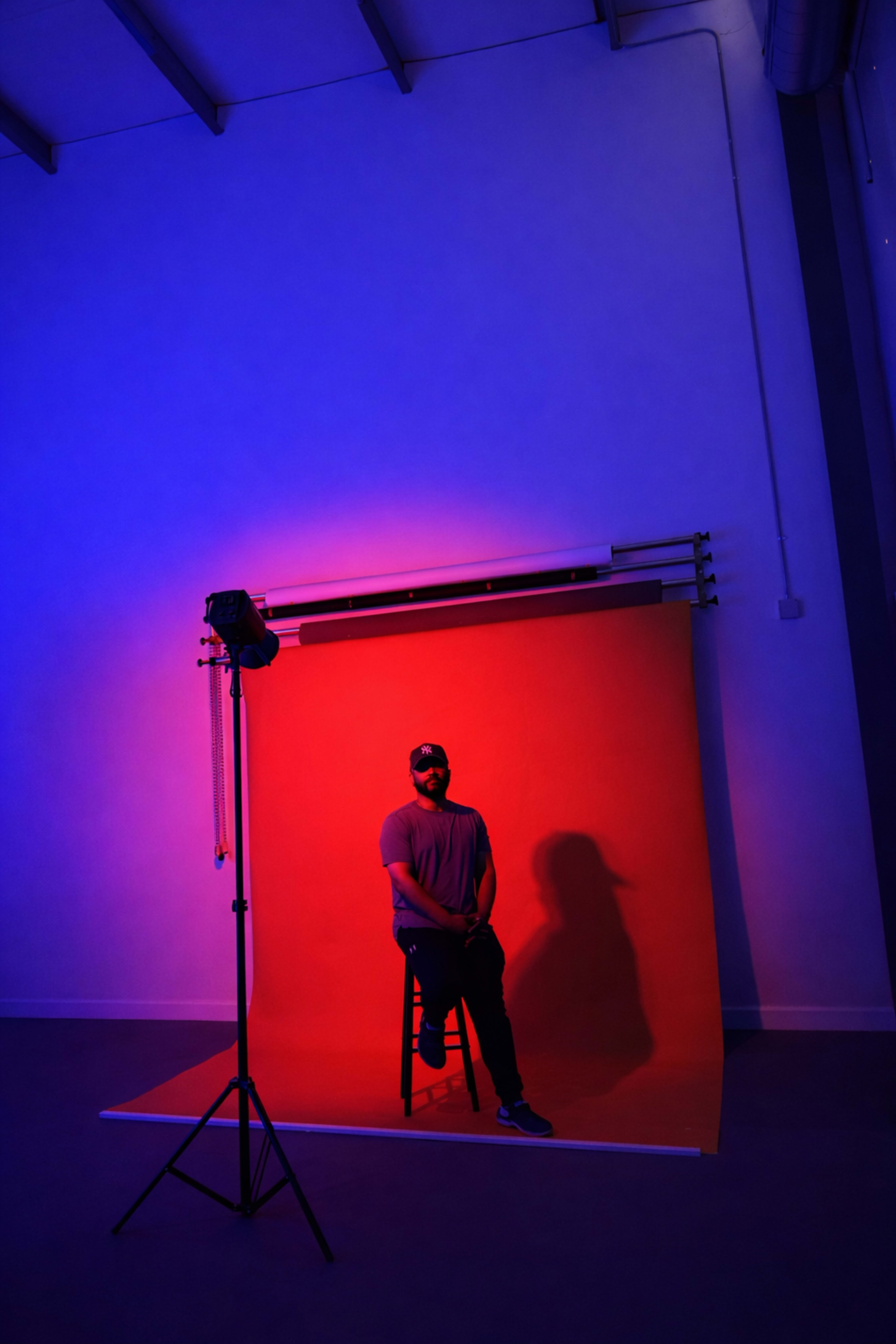 A man sits on a stool in front of a brightly colored backdrop illuminated by dramatic lighting.