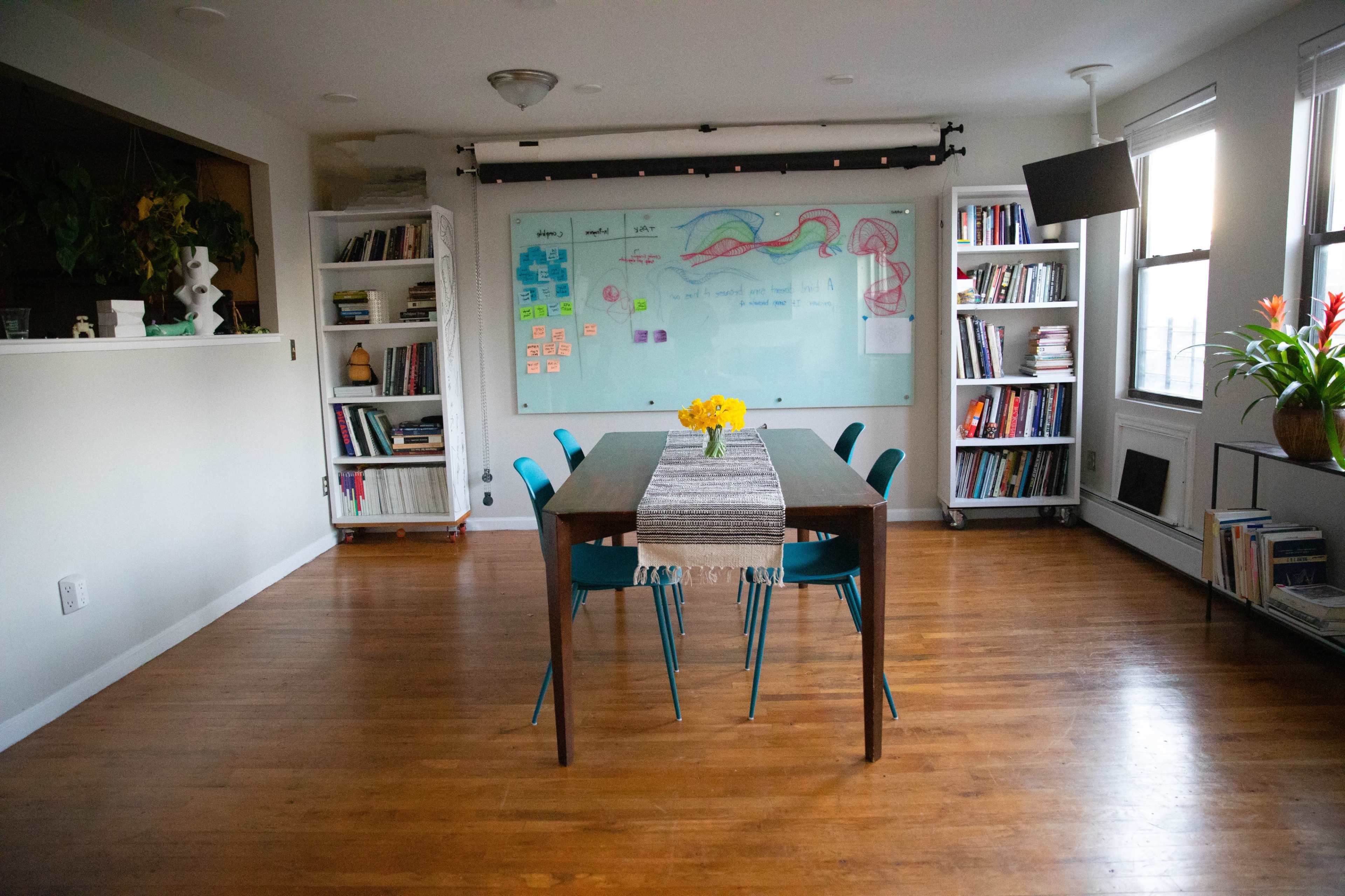 A dining area features a long table with a striped runner and blue chairs, surrounded by shelves filled with books and a large whiteboard on the wall.