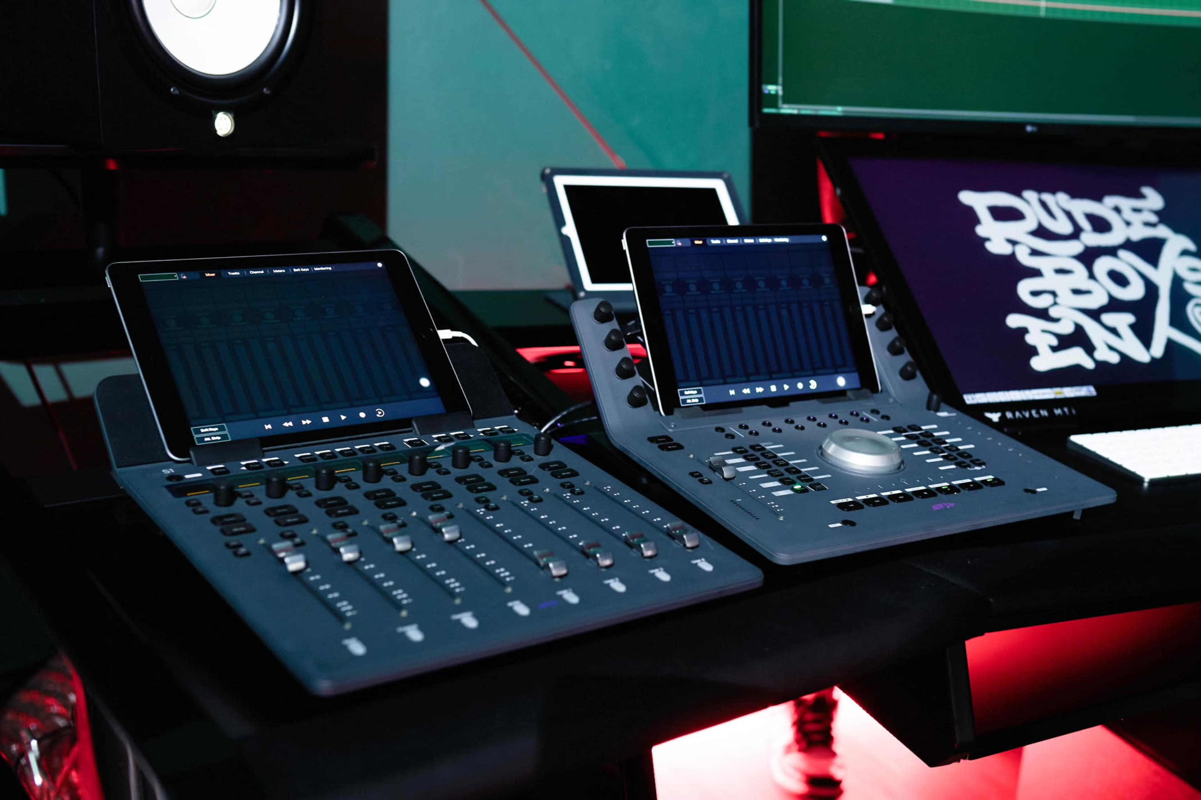 The image shows a modern music production setup featuring two digital mixing consoles and an audio interface on a desk, with speakers and a monitor in the background.