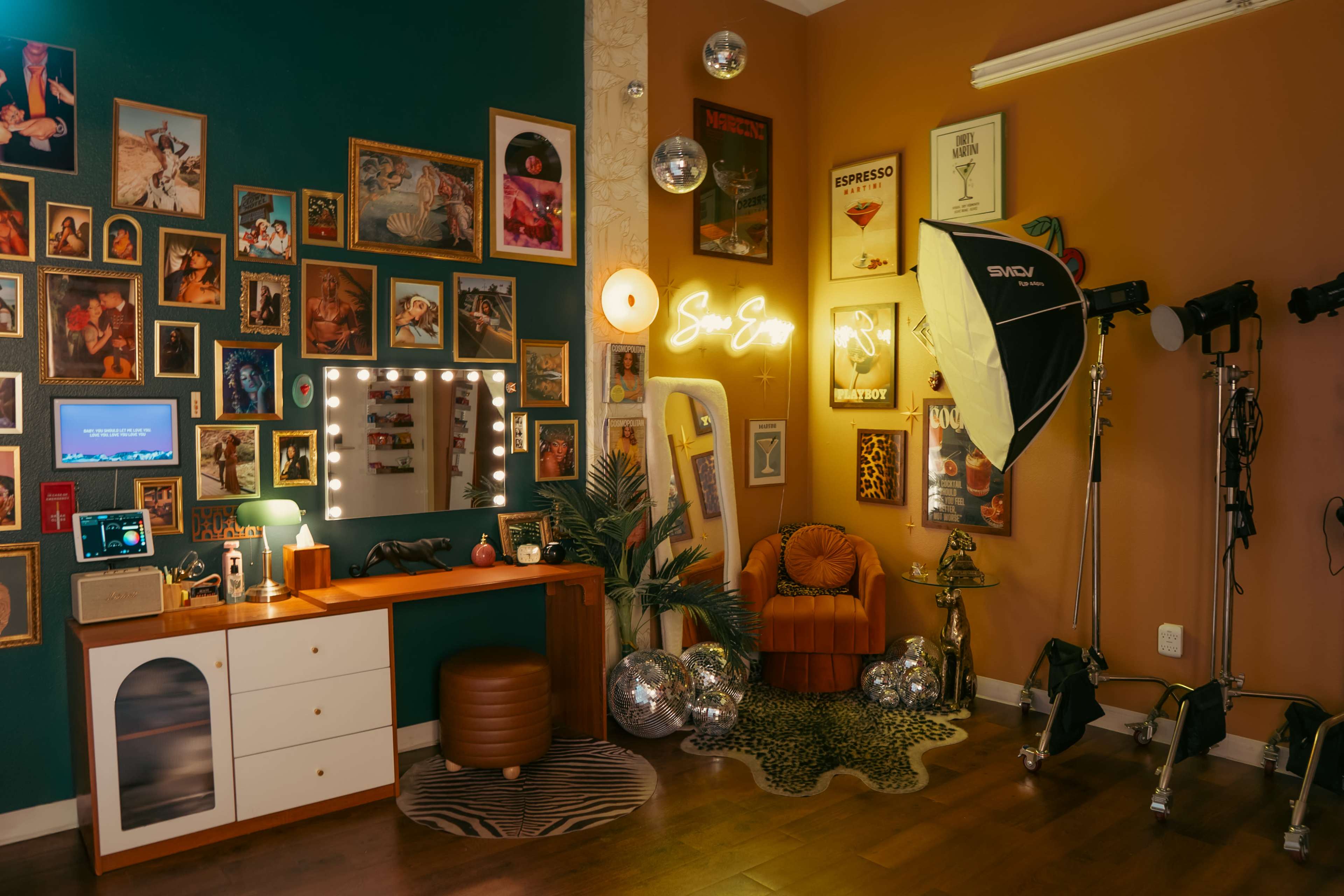 The image shows a stylish interior space featuring a vanity area with a mirror surrounded by lights, a comfortable chair, framed photos on the walls, and a corner setup for photography with studio lighting.