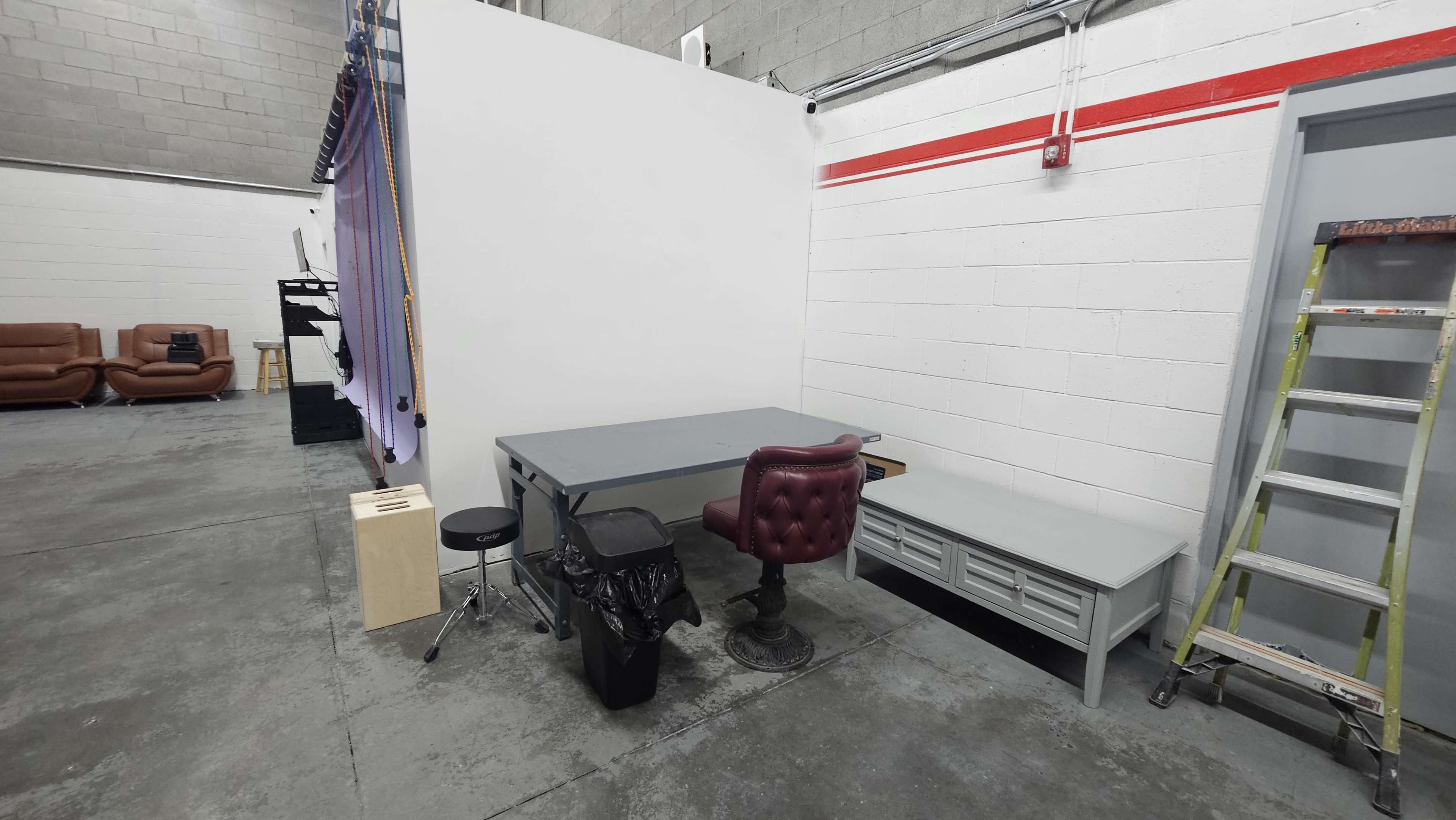 The image shows a small workspace in a warehouse, featuring a gray desk, a maroon leather chair, a stool, a trash can, and a bench against a white wall.