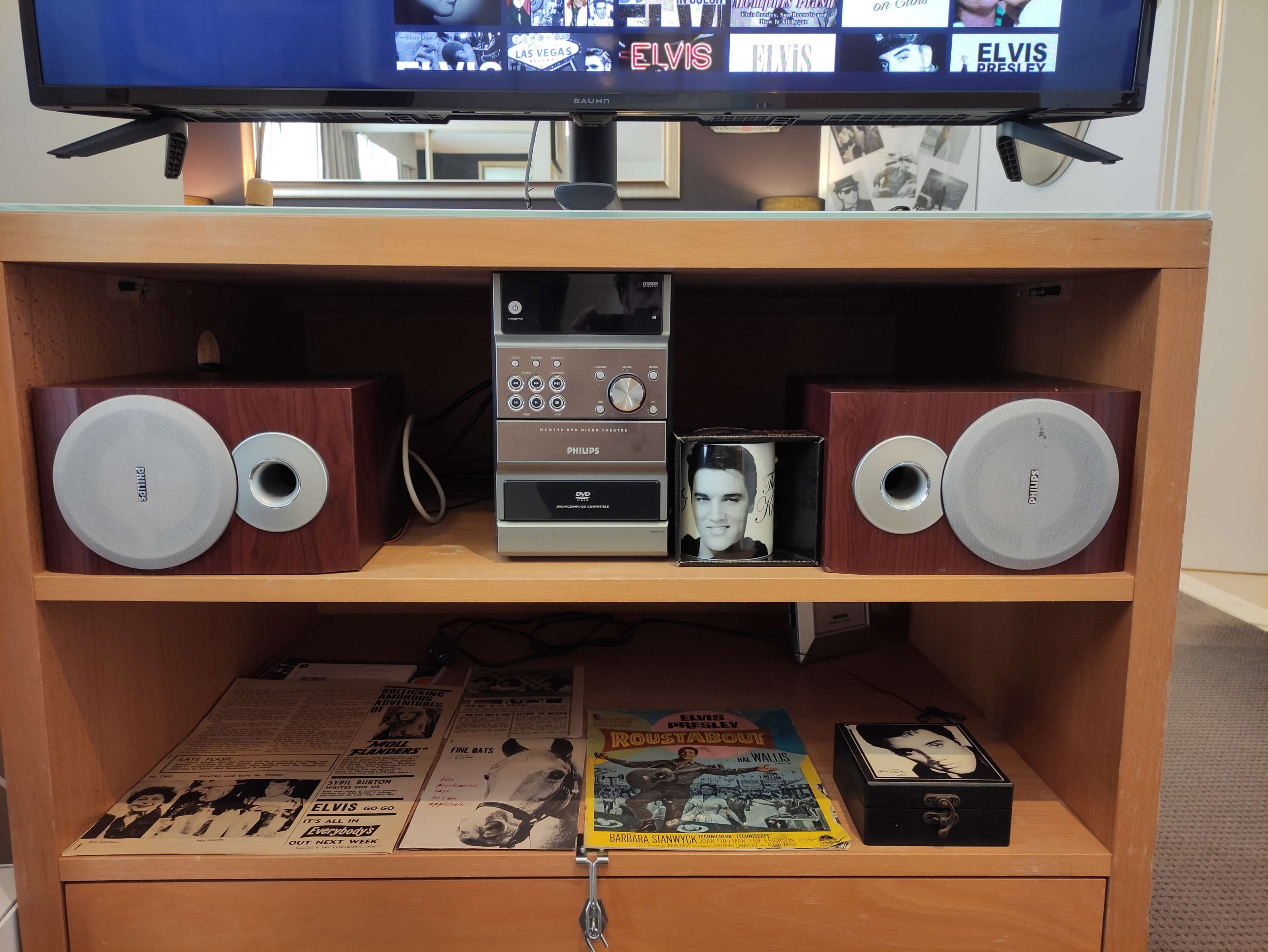 A wooden media console with a Philips CD player and two speakers, alongside various Elvis memorabilia and magazines.