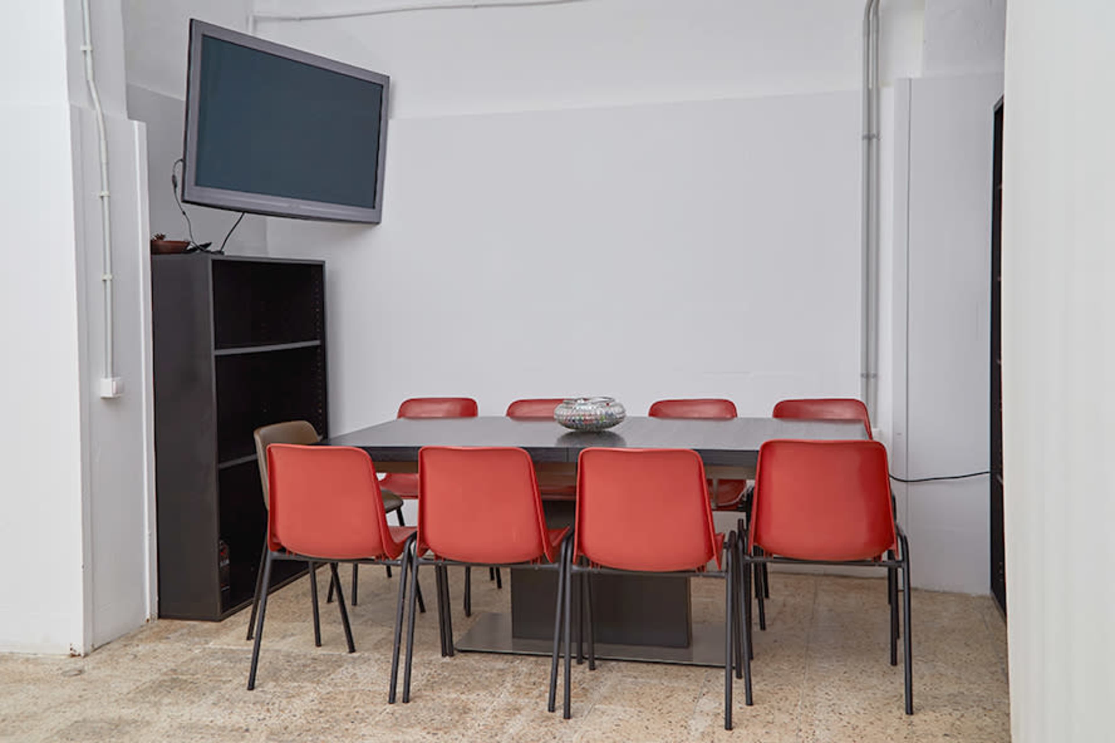 A rectangular meeting table surrounded by red chairs is positioned in a room with a mounted television and a shelving unit.