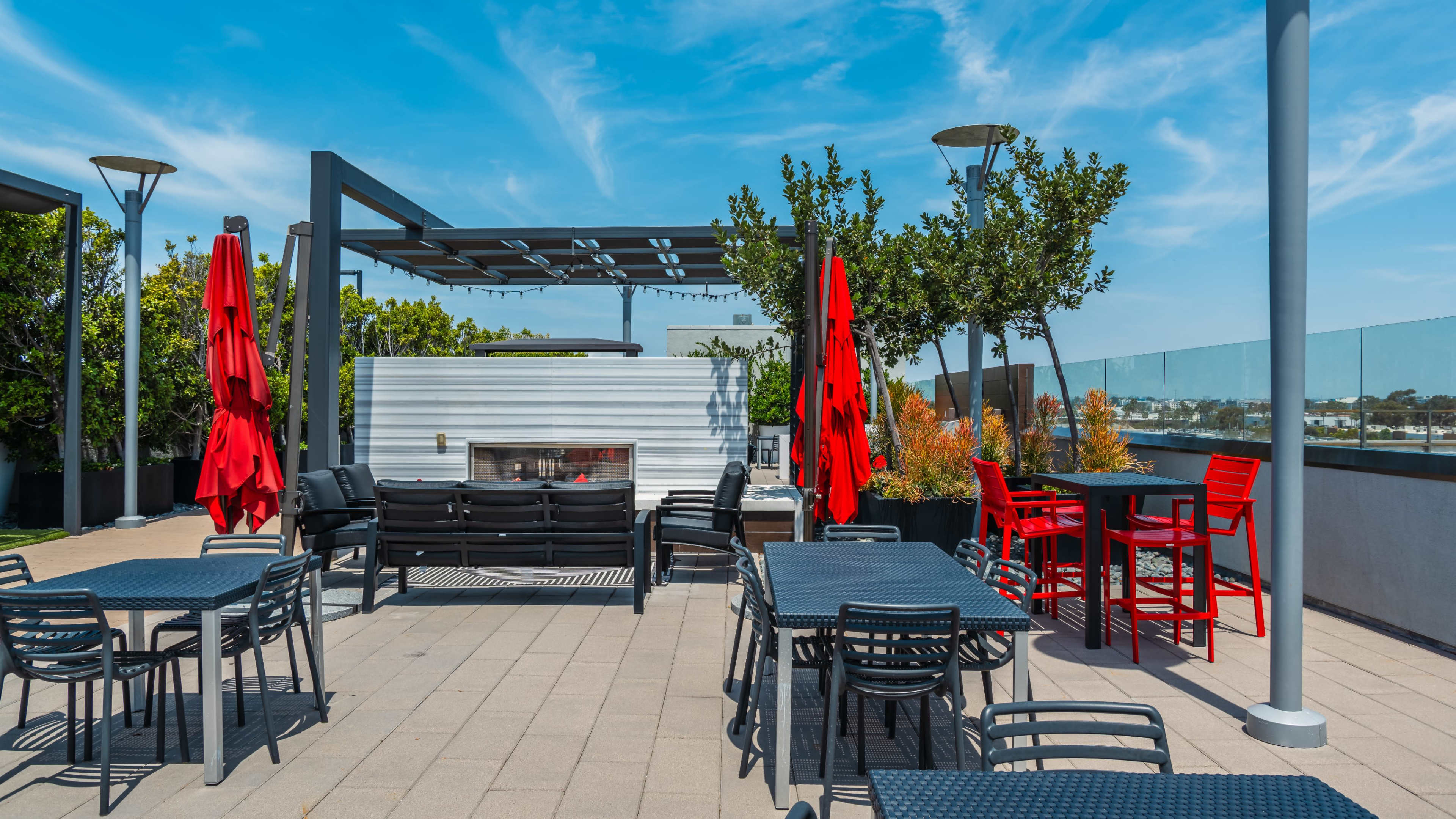 The image shows a spacious outdoor terrace featuring several black tables and chairs, red umbrella seating, and a white structure with a fireplace, all under a clear blue sky.