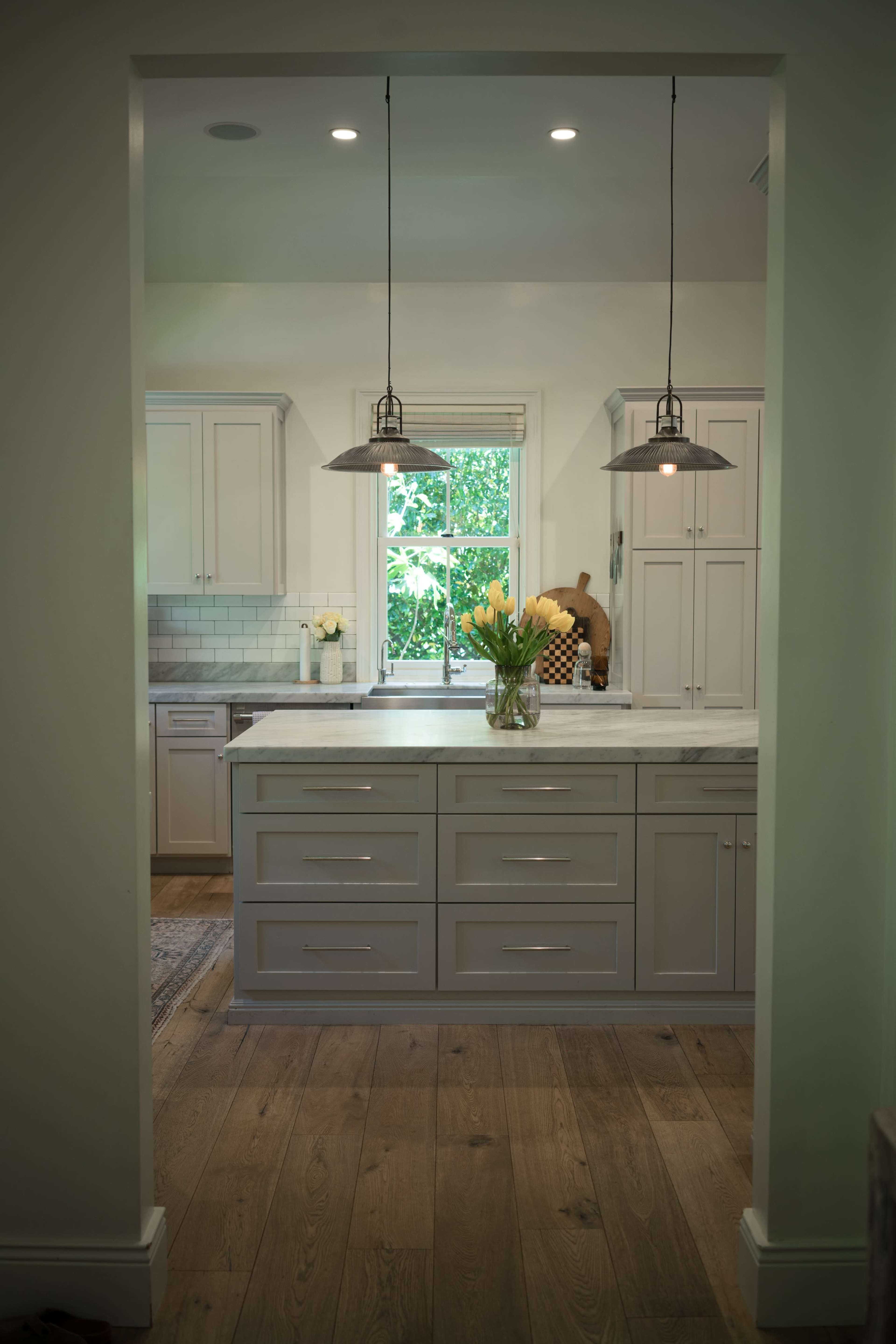 A well-lit kitchen features gray cabinetry, a large marble countertop island, and a vase of yellow flowers.