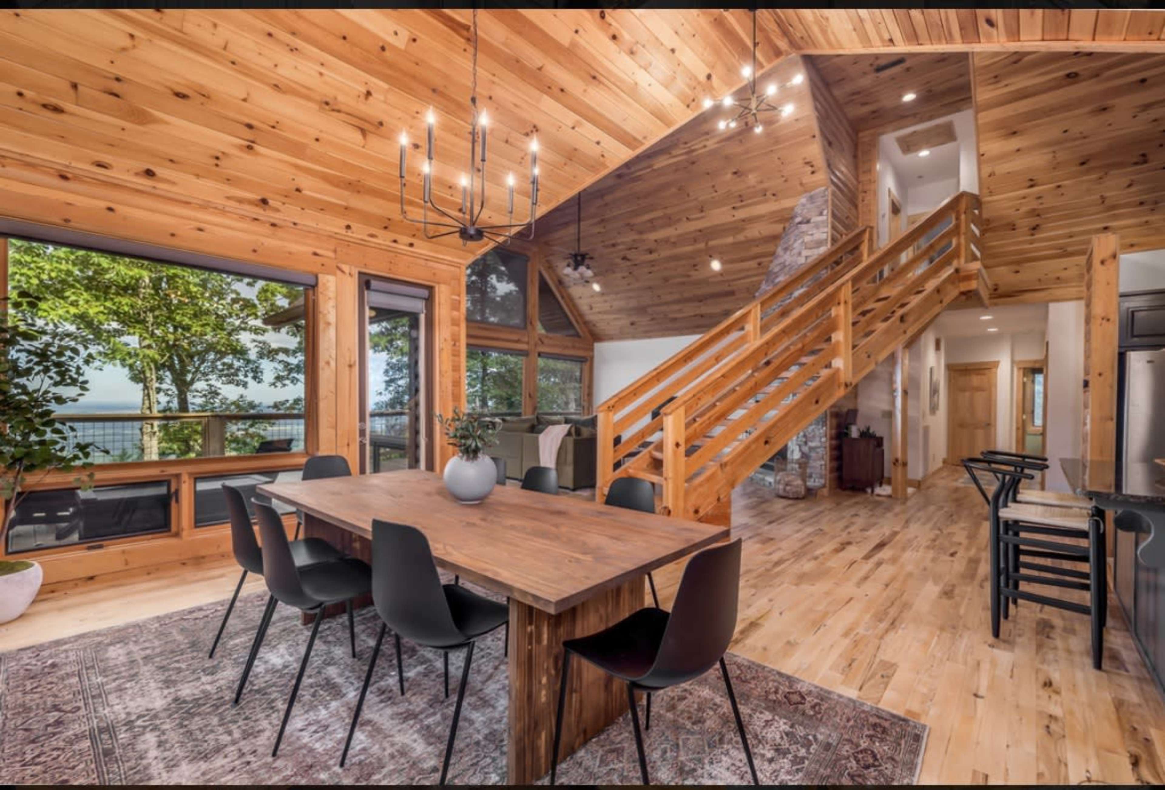 A wooden dining area features a large table surrounded by black chairs, with large windows offering a view of trees and a staircase leading to an upper level.