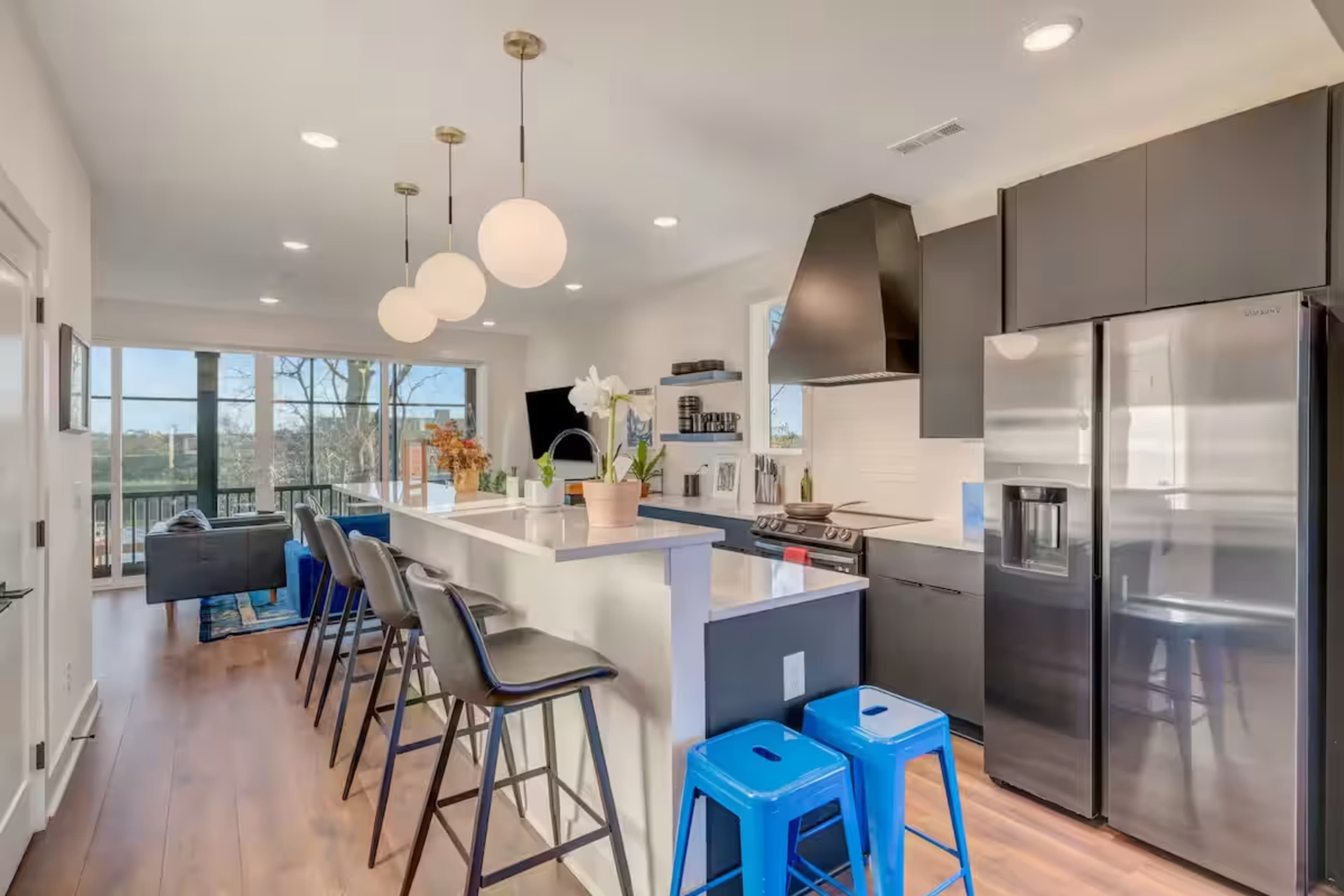 The image shows a modern kitchen with a large island, bar seating, sleek appliances, and large windows offering a view of the outdoors.