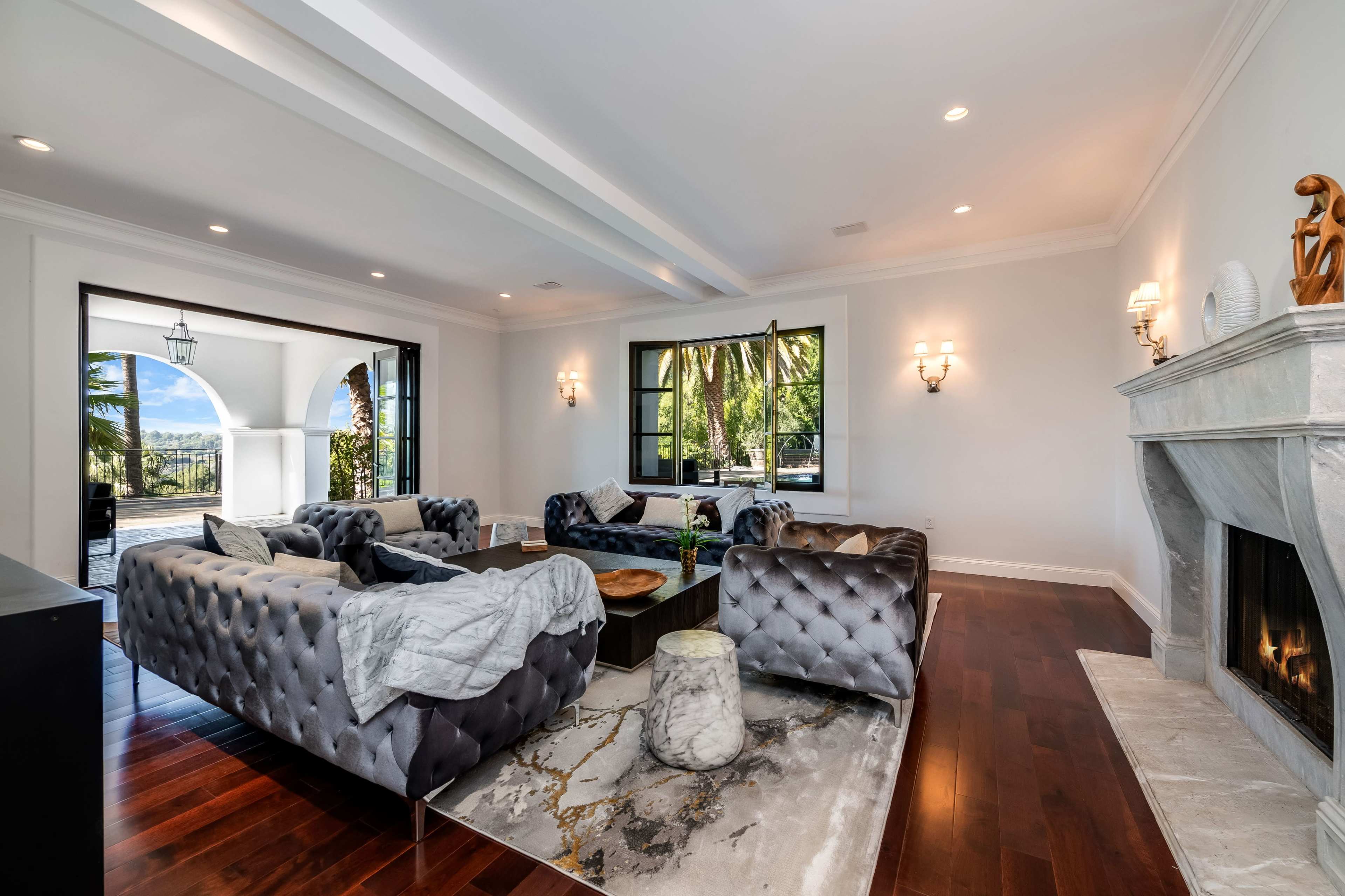 The image shows a spacious living room with tufted velvet sofas, a marble fireplace, and large windows framing a view of palm trees.
