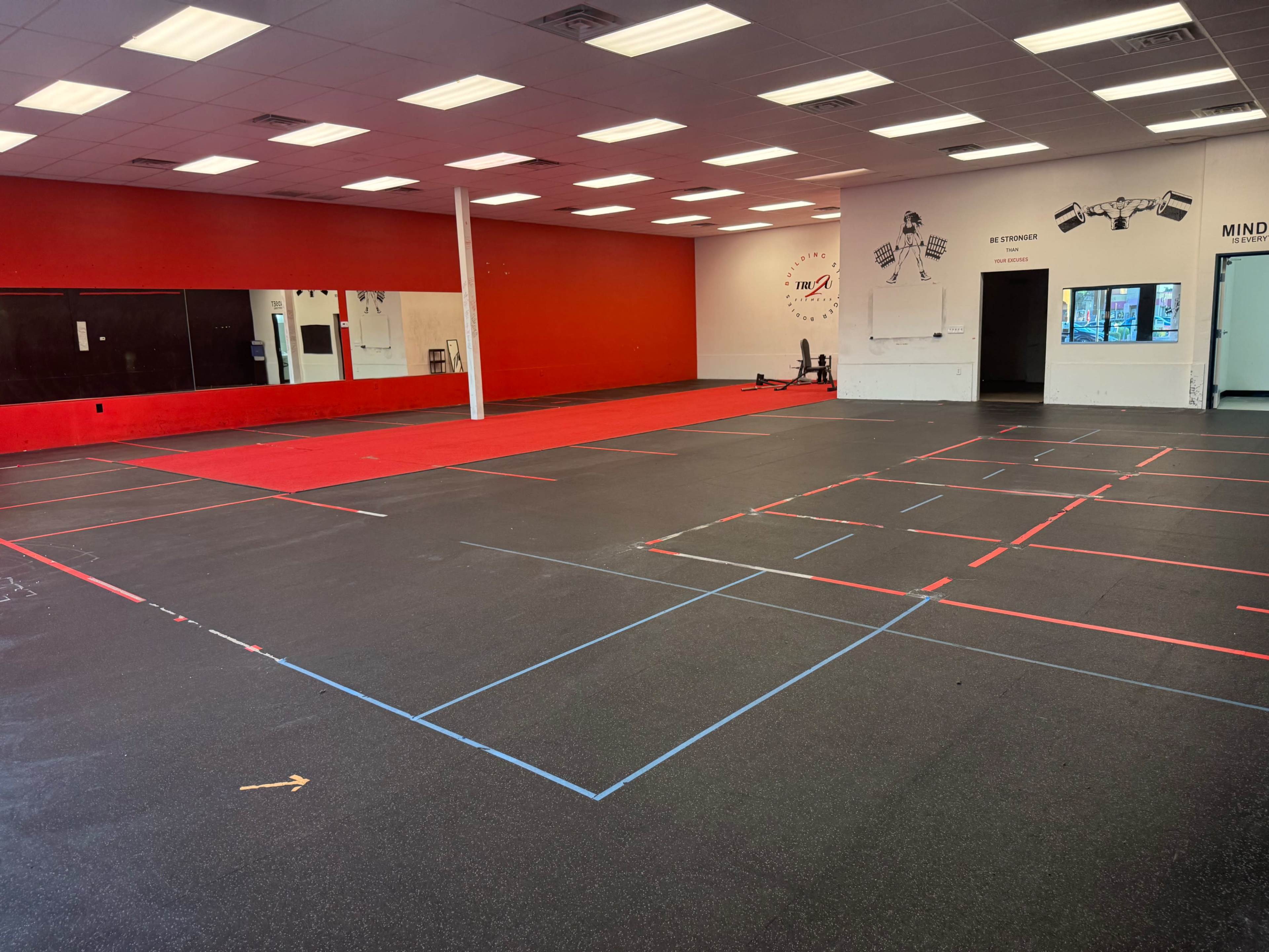 The image shows an empty gym space with black rubber flooring marked by red and blue tape lines and mirrors on one wall.