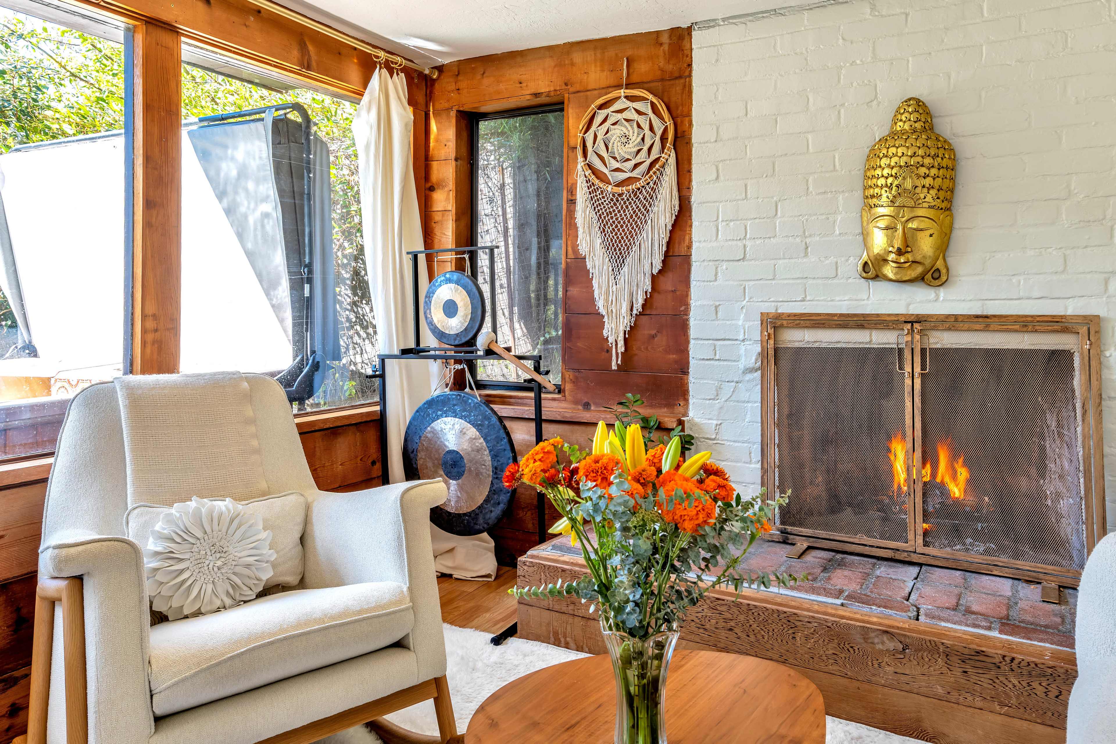 The image shows a cozy living room with a comfortable armchair, a vase of flowers on a table, a warm fireplace, and decorative items on the walls and shelves.