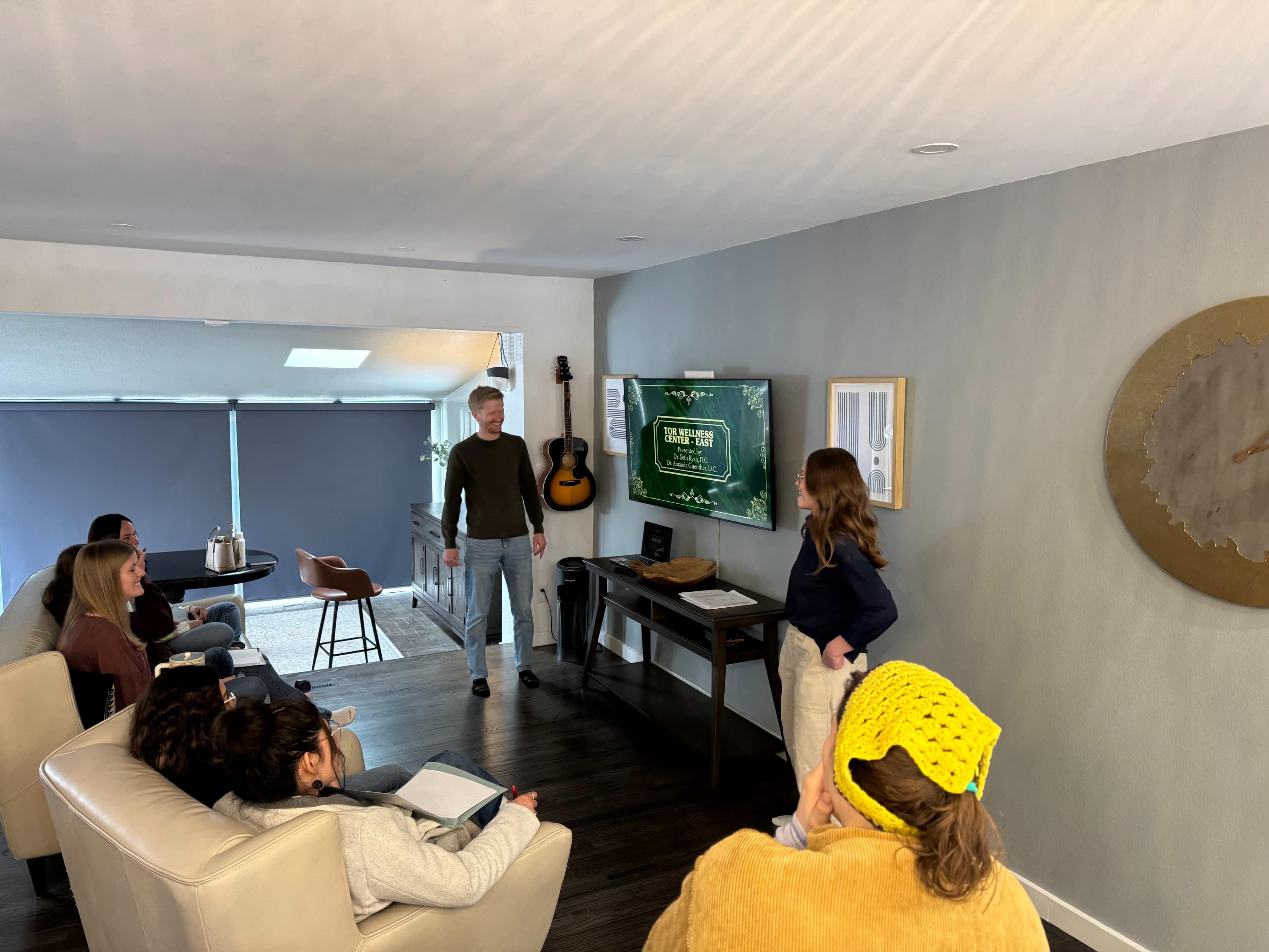 A group of six people is gathered in a living room, with one person standing and presenting in front of a television displaying a presentation.