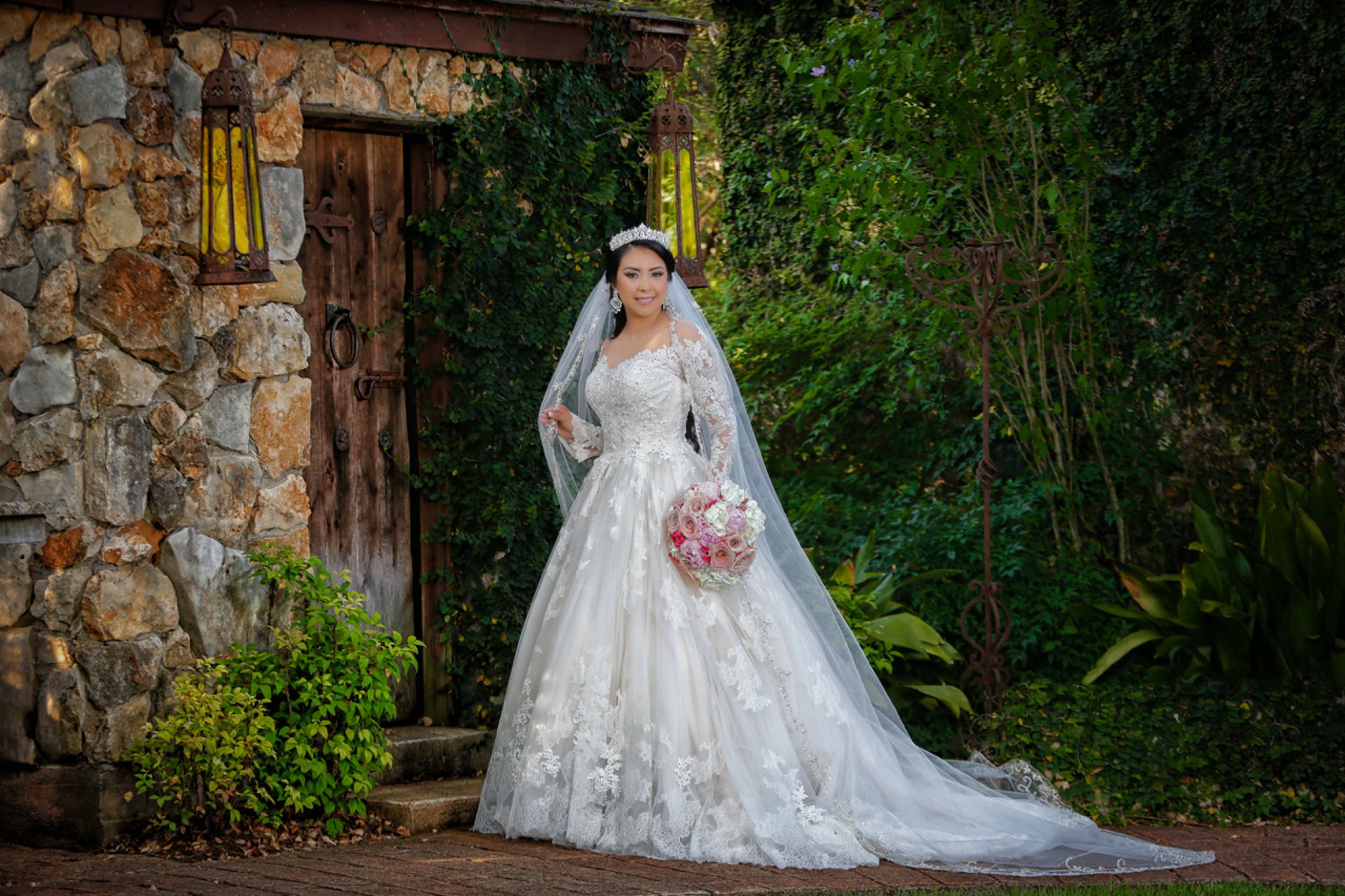 A bride in a white wedding dress stands outside a stone structure, holding a bouquet of flowers.