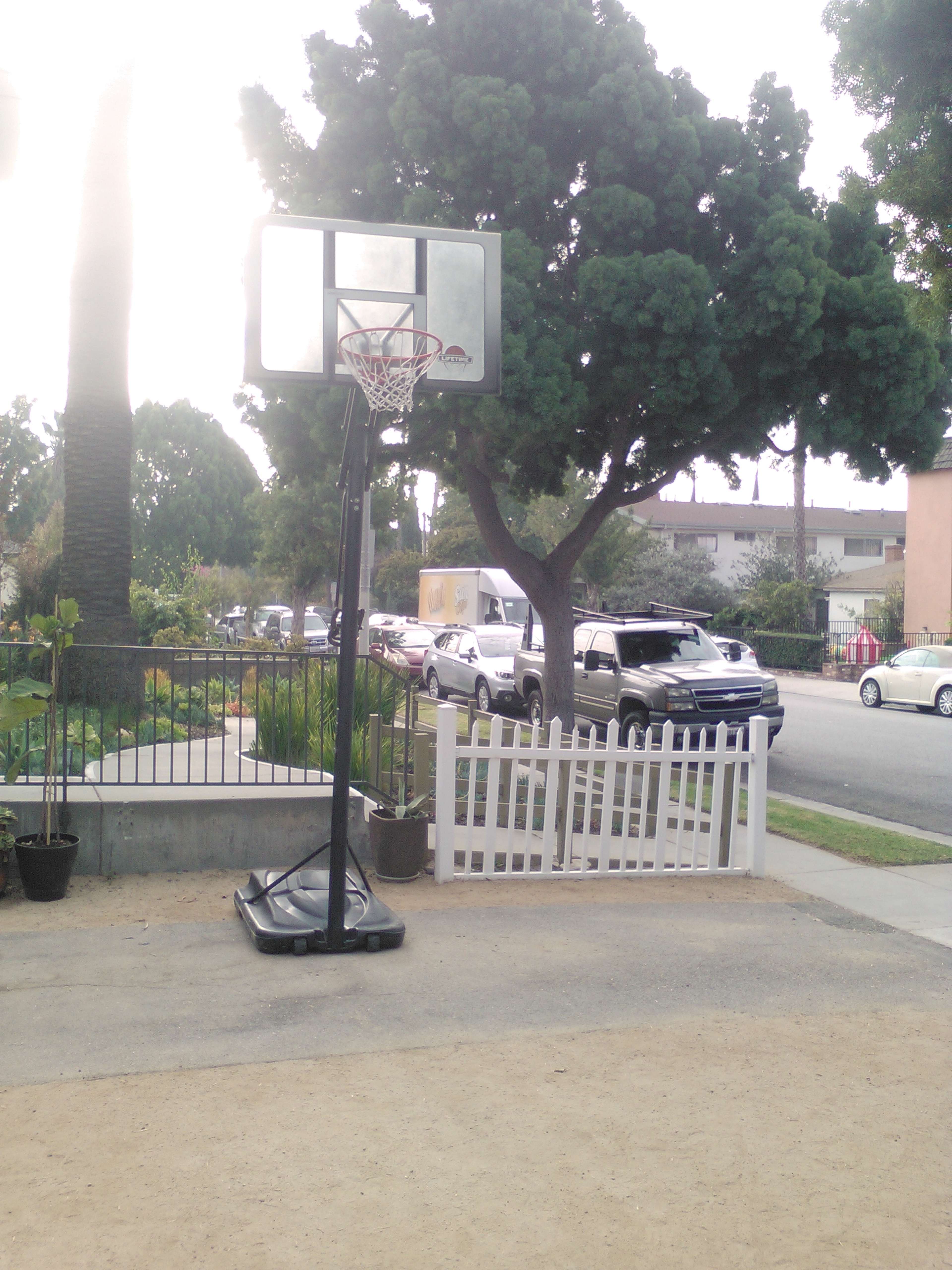 A basketball hoop stands on a gravel surface near a white picket fence, with parked cars and trees in the background.