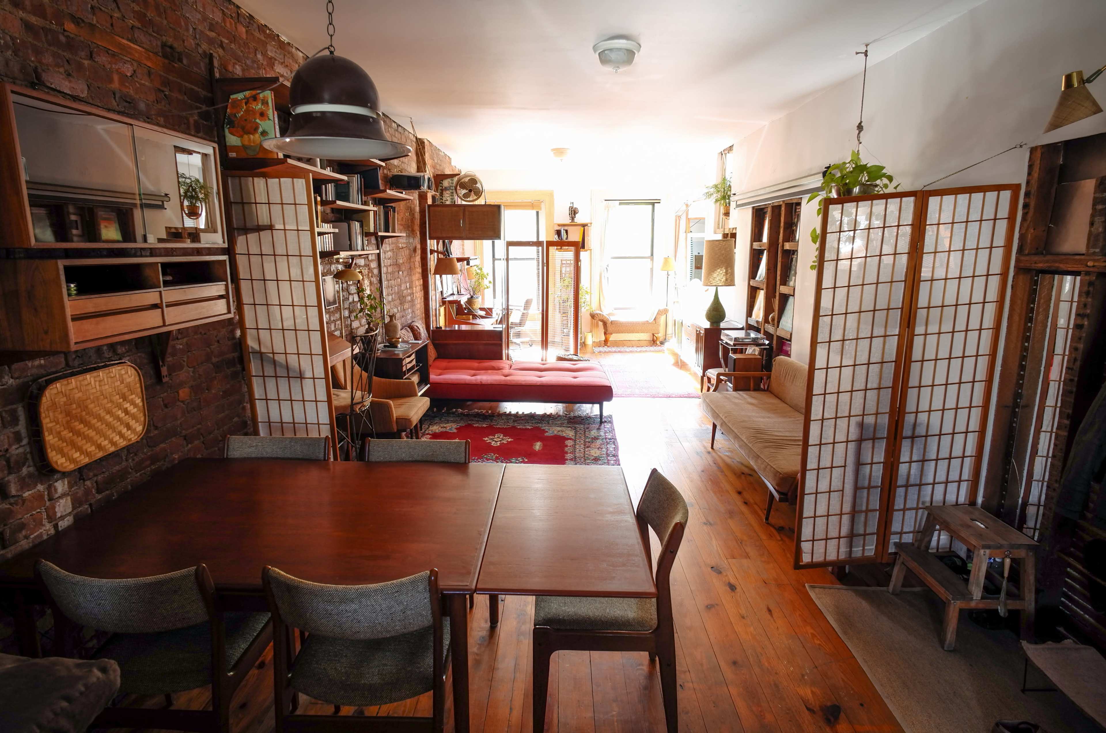 The image shows a cozy living space with wooden furniture, divided by shoji screens, featuring a dining area and a seating area filled with plants and natural light.