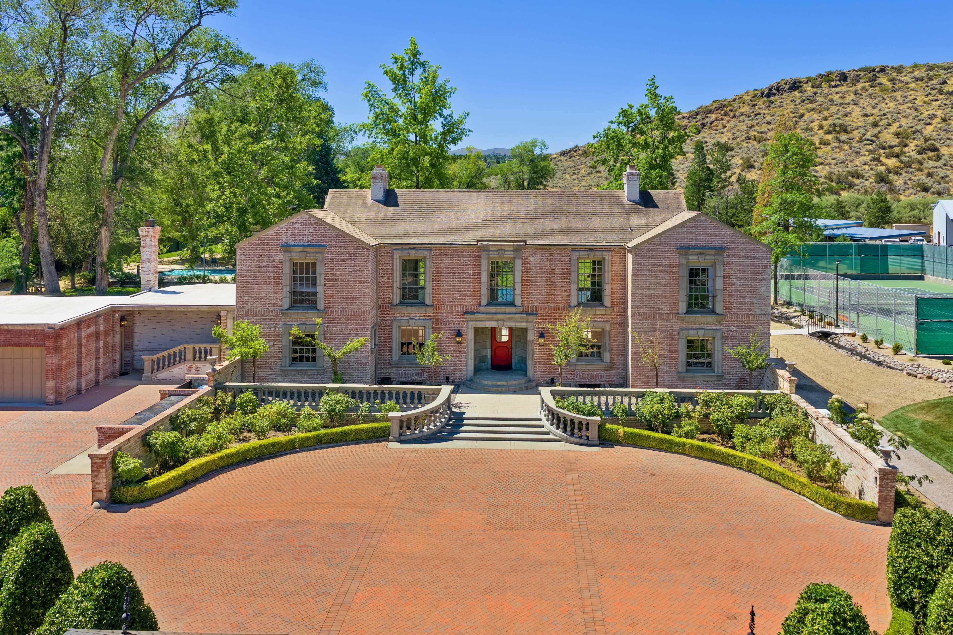 A large brick house with a symmetrical facade and a landscaped front yard is set against a backdrop of trees and hills.