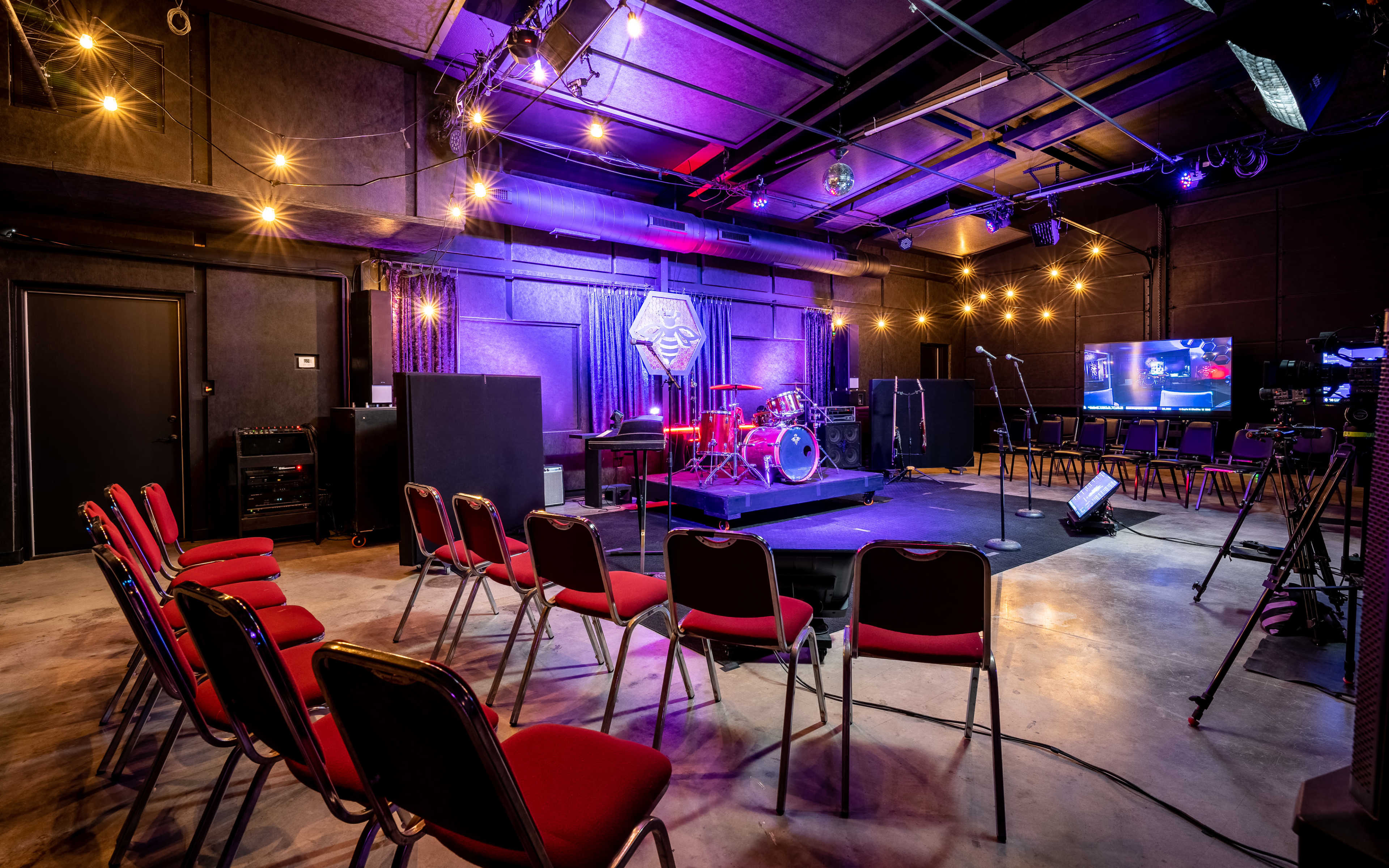 The image shows a stage setup in a music venue with drum equipment, sound gear, and rows of red chairs arranged for an audience.