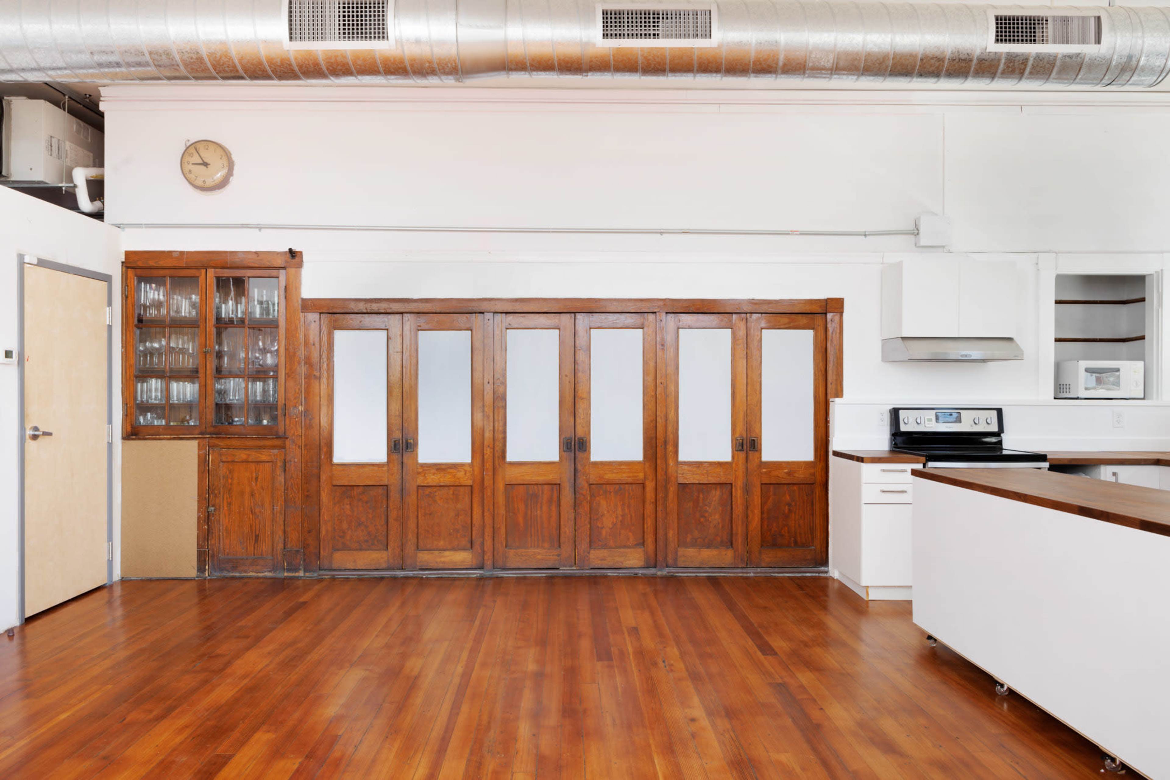 The image shows a spacious kitchen area featuring a wooden cabinet with glass doors, multiple wooden panel doors, and an adjacent modern kitchen counter with appliances against a backdrop of hardwood flooring.