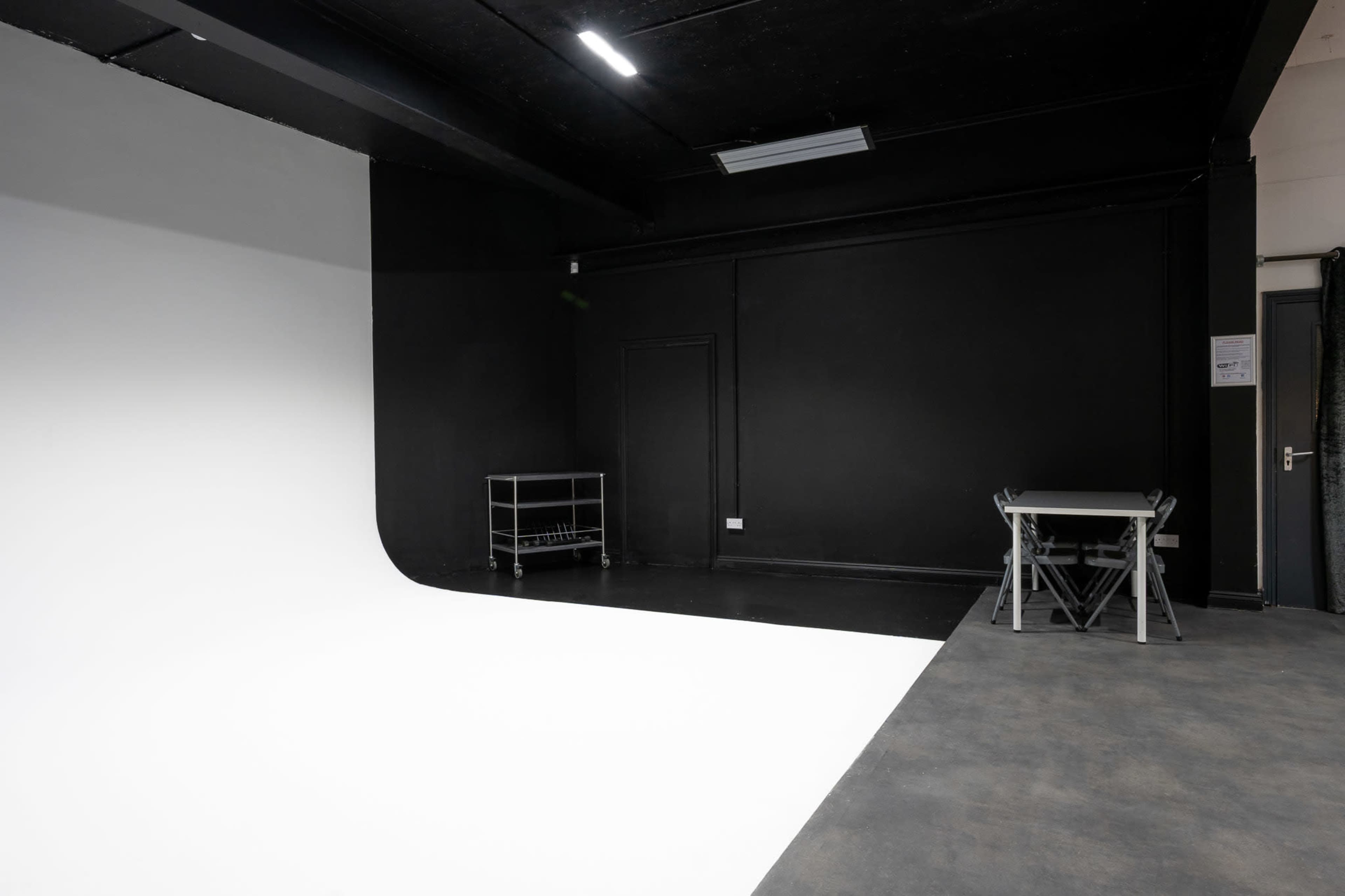The image shows a photography studio featuring a white backdrop, black walls, and a small table with chairs in one corner.