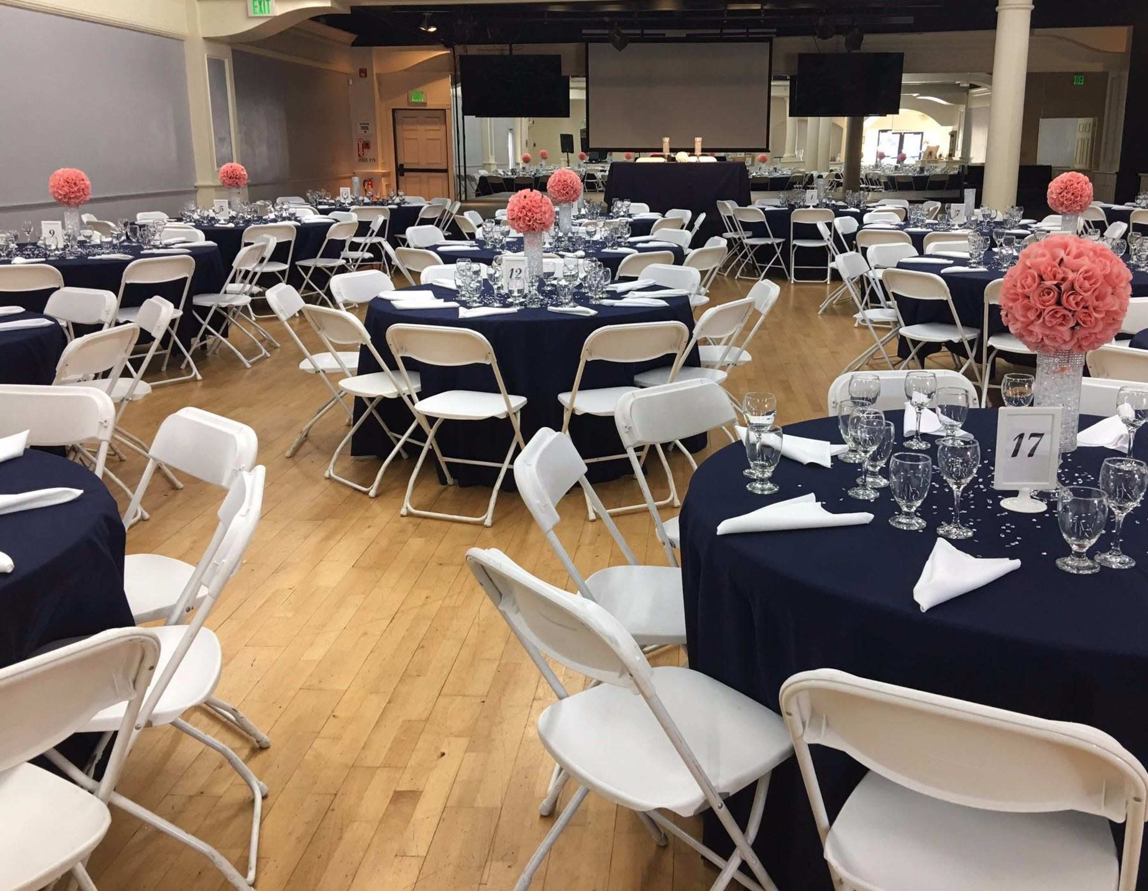 The image shows a banquet hall set up for an event, featuring round tables draped in navy tablecloths adorned with pink floral centerpieces and surrounded by white folding chairs.