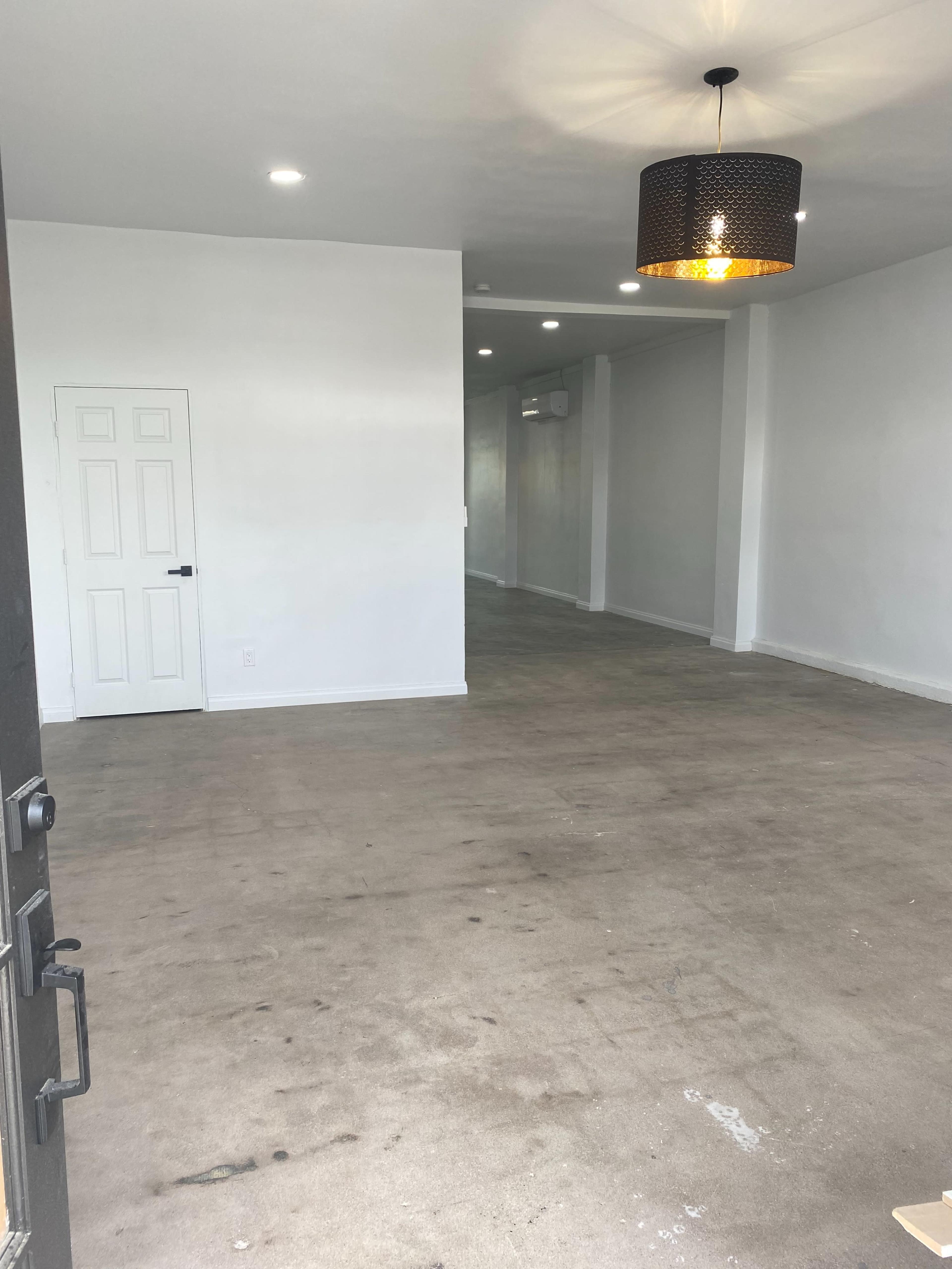 The image shows a vacant room with light-colored walls, a concrete floor, and a modern pendant light hanging from the ceiling.