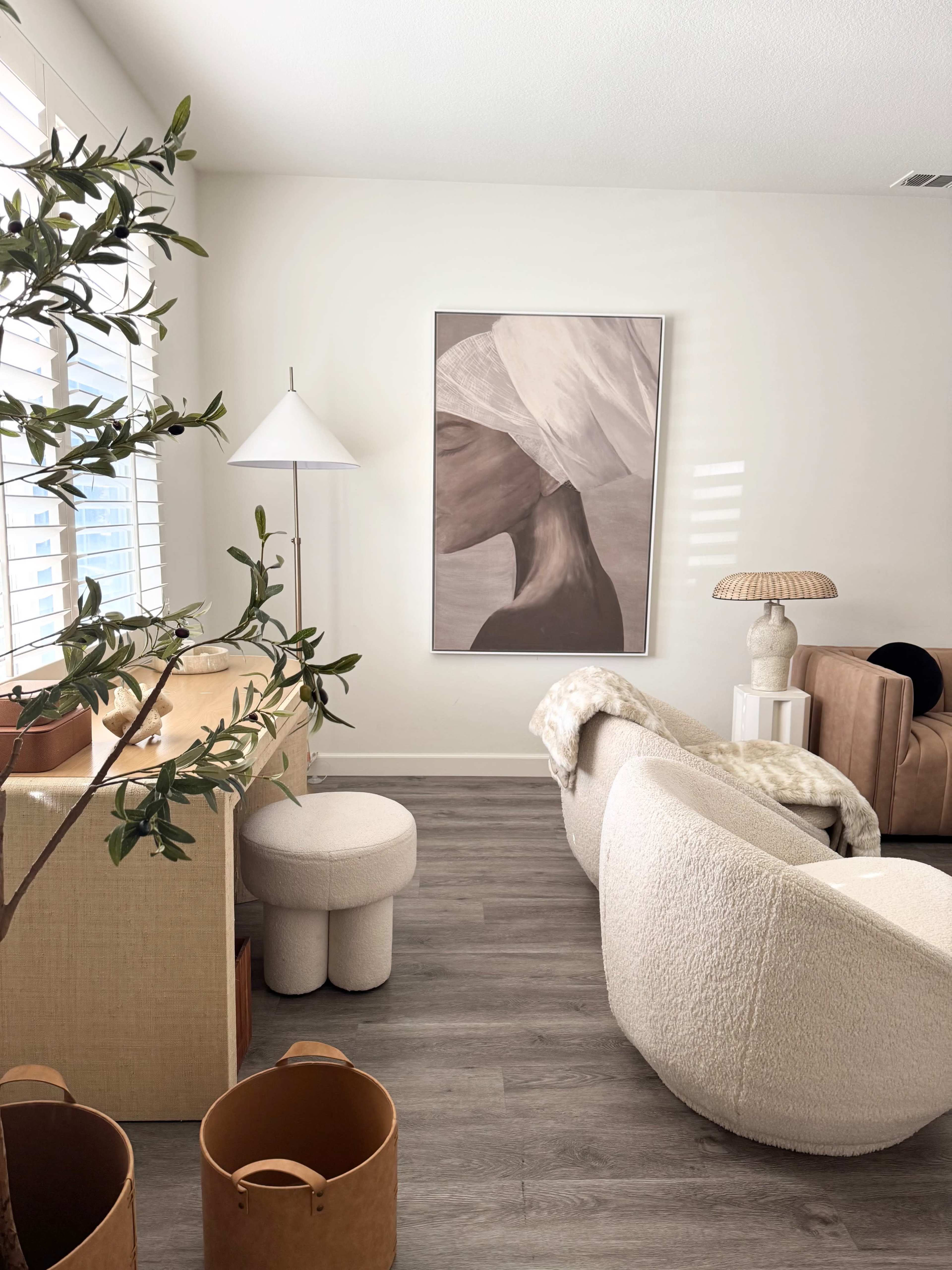 The image shows a modern living room featuring a large artwork of a profile, a lamp, and various seating options, with natural light streaming in through the windows.