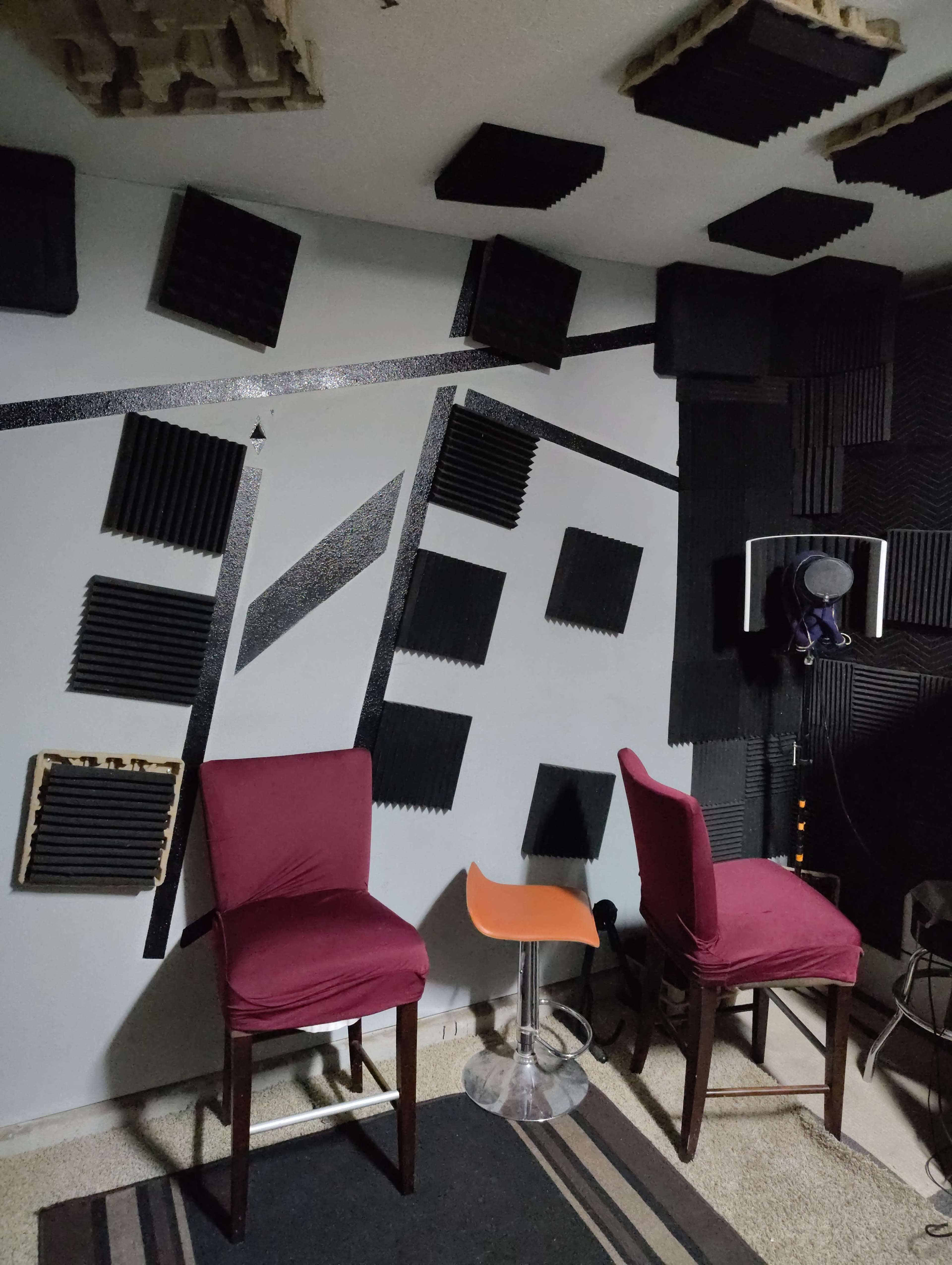 The image shows a soundproofed recording studio with two chairs, one wooden and one transparent, set against walls lined with acoustic foam panels.