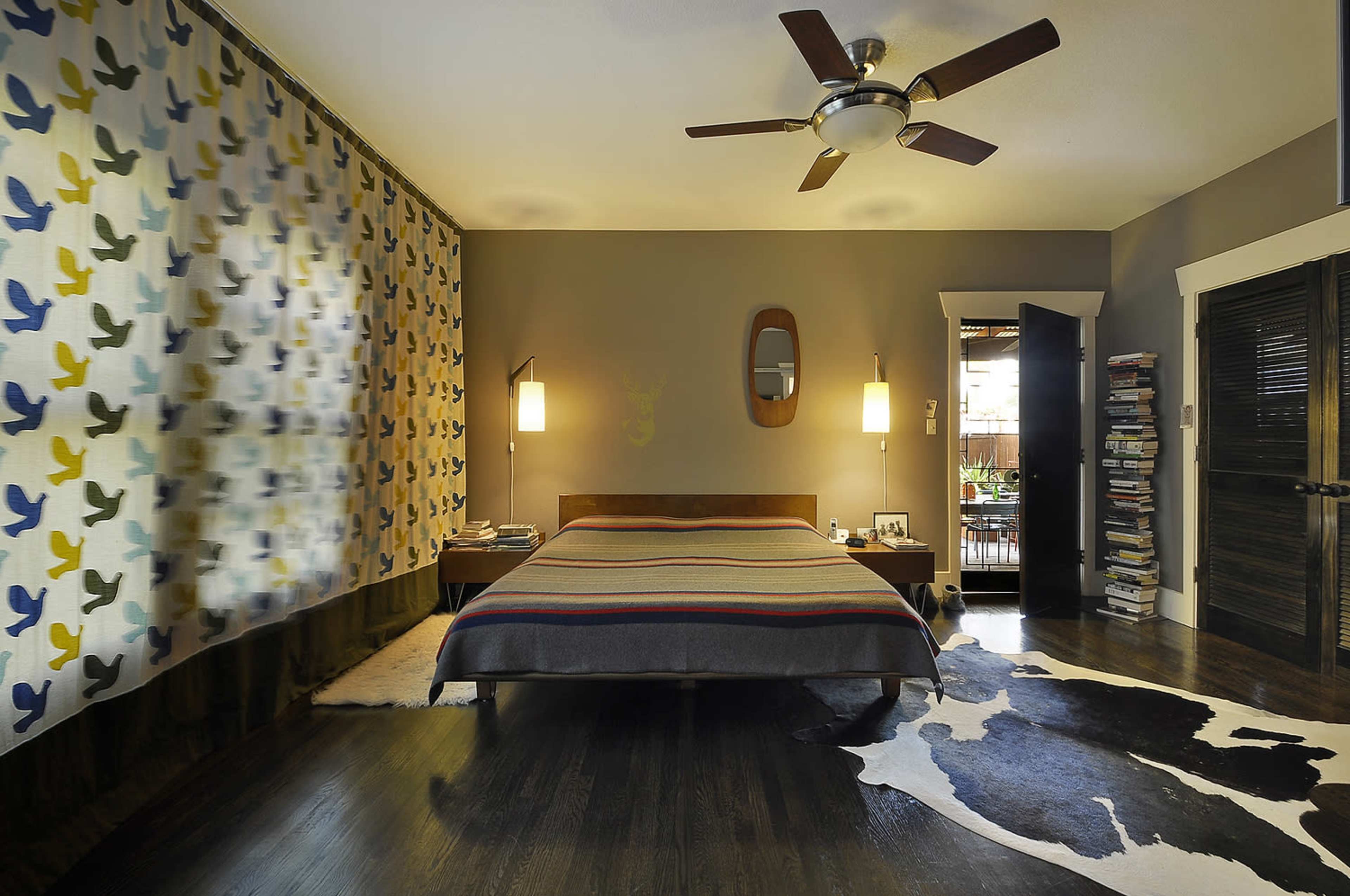 The image features a modern bedroom with a wooden bed, a patterned curtain, a cowhide rug, and a stack of books beside a doorway.