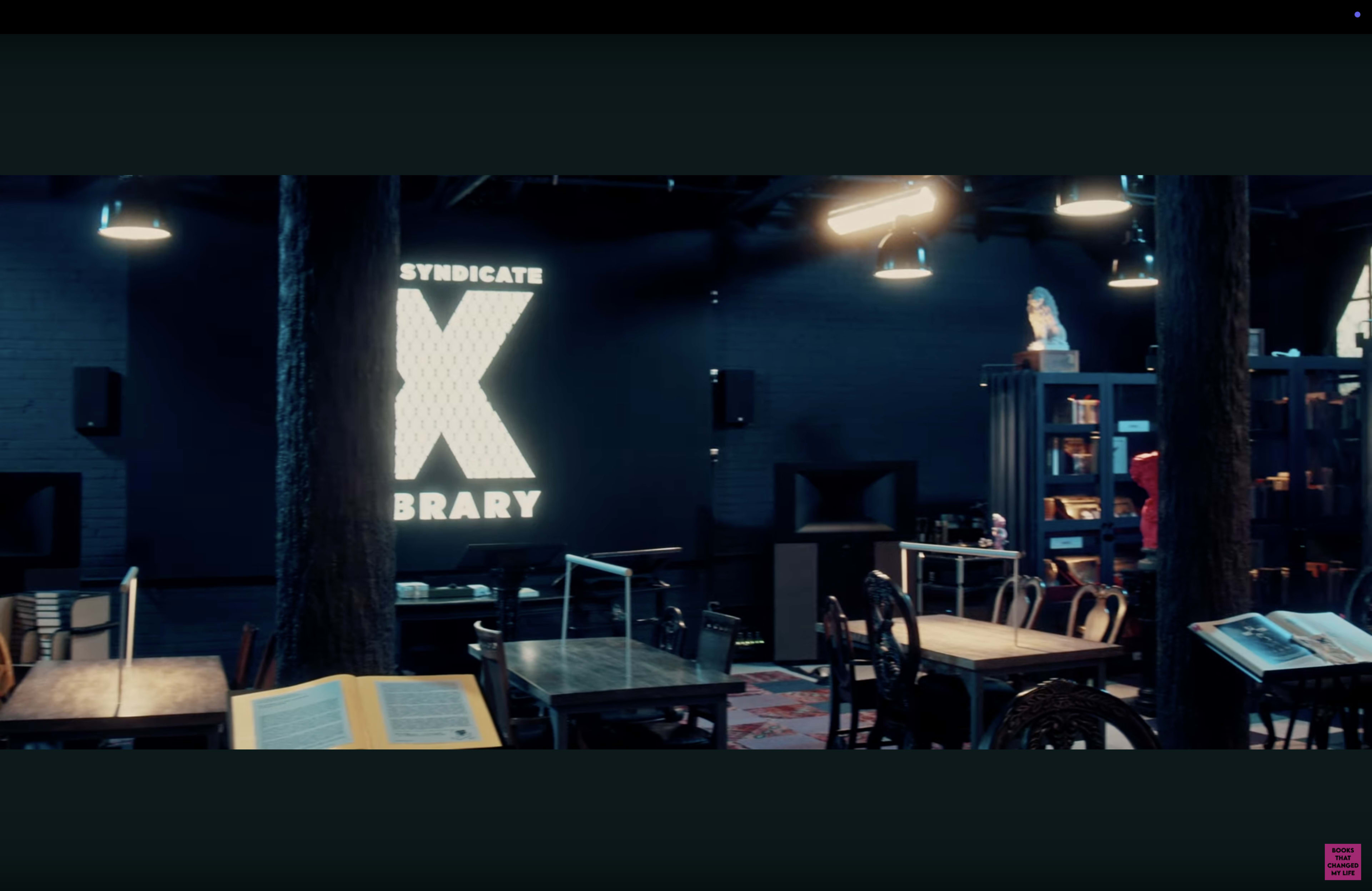 The image shows a modern café or library interior with wooden tables and chairs, featuring a large illuminated sign that reads "SYNDICATE X BRARY" on the wall.