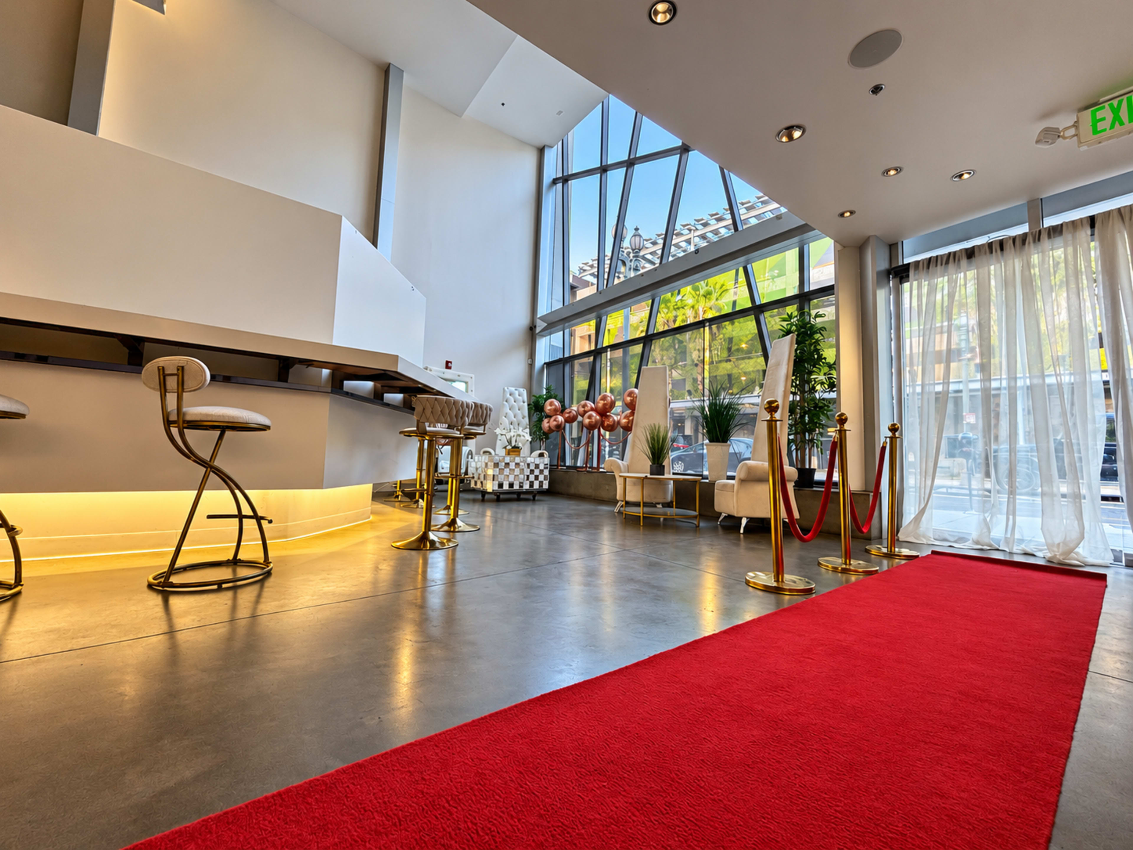 The image shows a modern lobby featuring a red carpet, bar stools, plants, and large windows letting in natural light.
