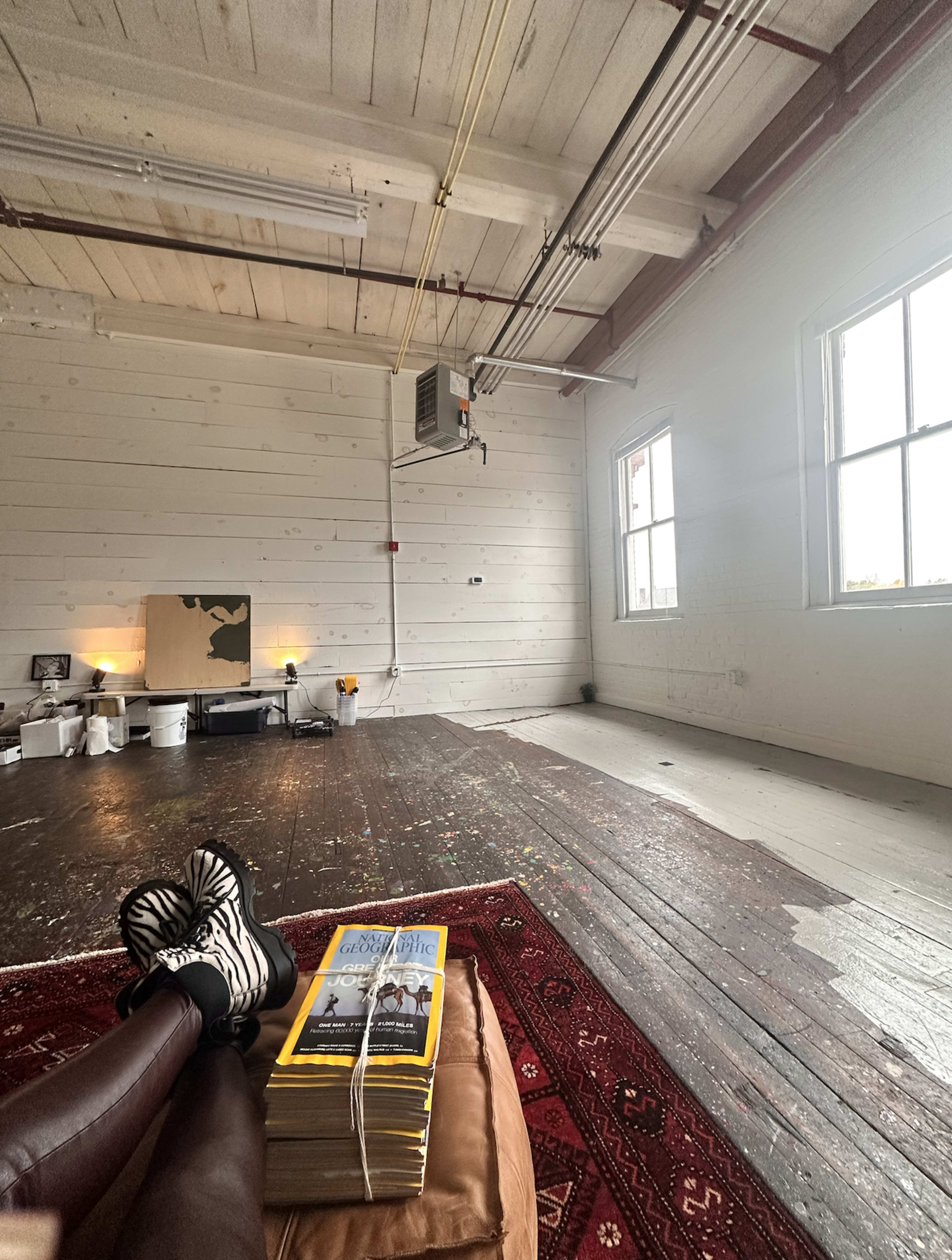 The image shows a spacious, mostly empty room with wooden flooring, featuring a stack of magazines on a large cushion and two windows allowing natural light to enter.