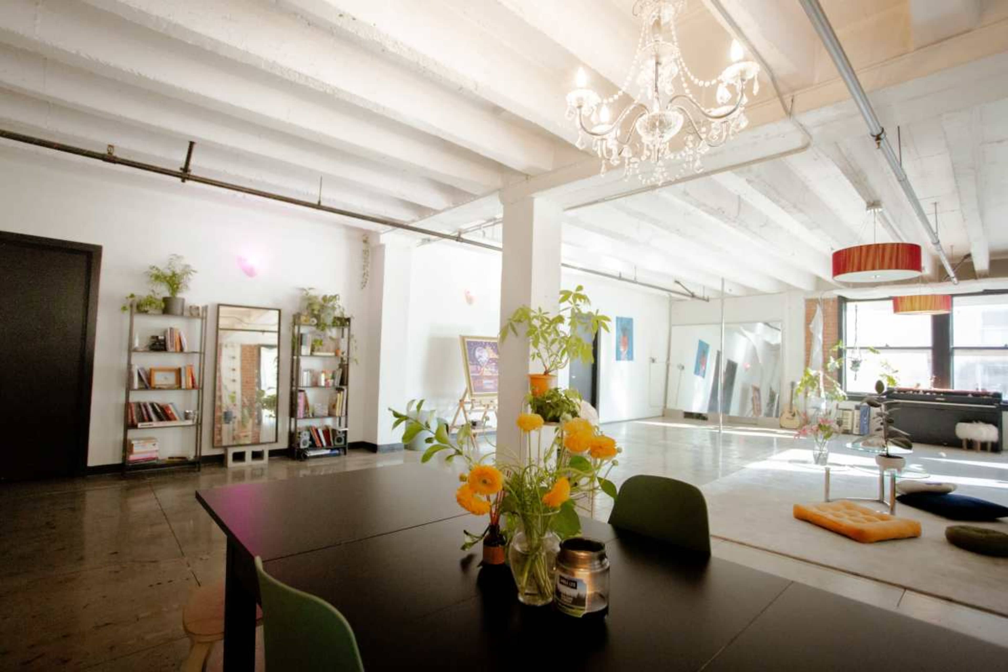 A spacious, well-lit room features a dining table with a vase of flowers, bookshelves, and various artworks displayed on the walls.