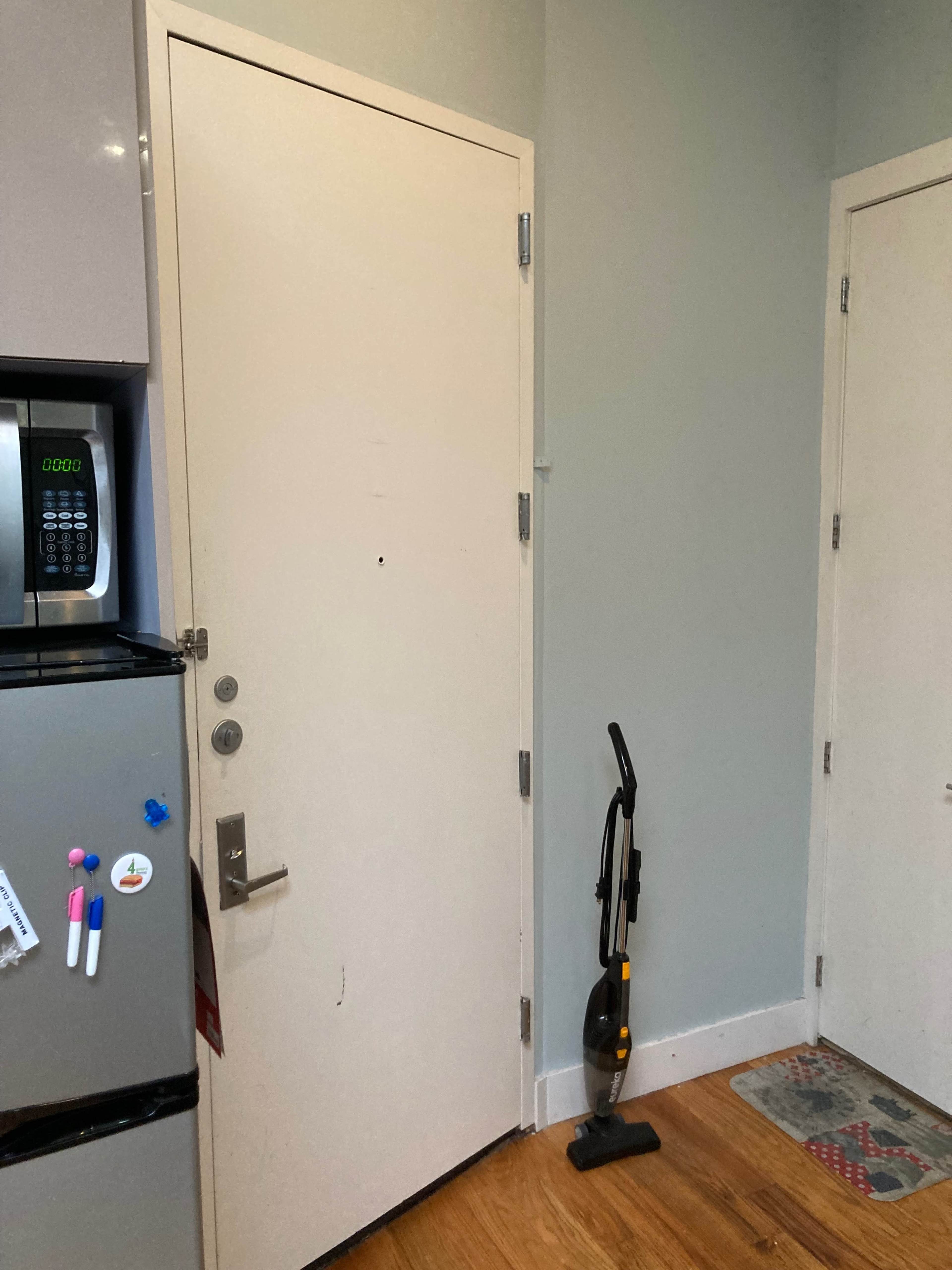 A vacuum cleaner stands upright against a wall near a closed door in a kitchen area with a refrigerator and a microwave.