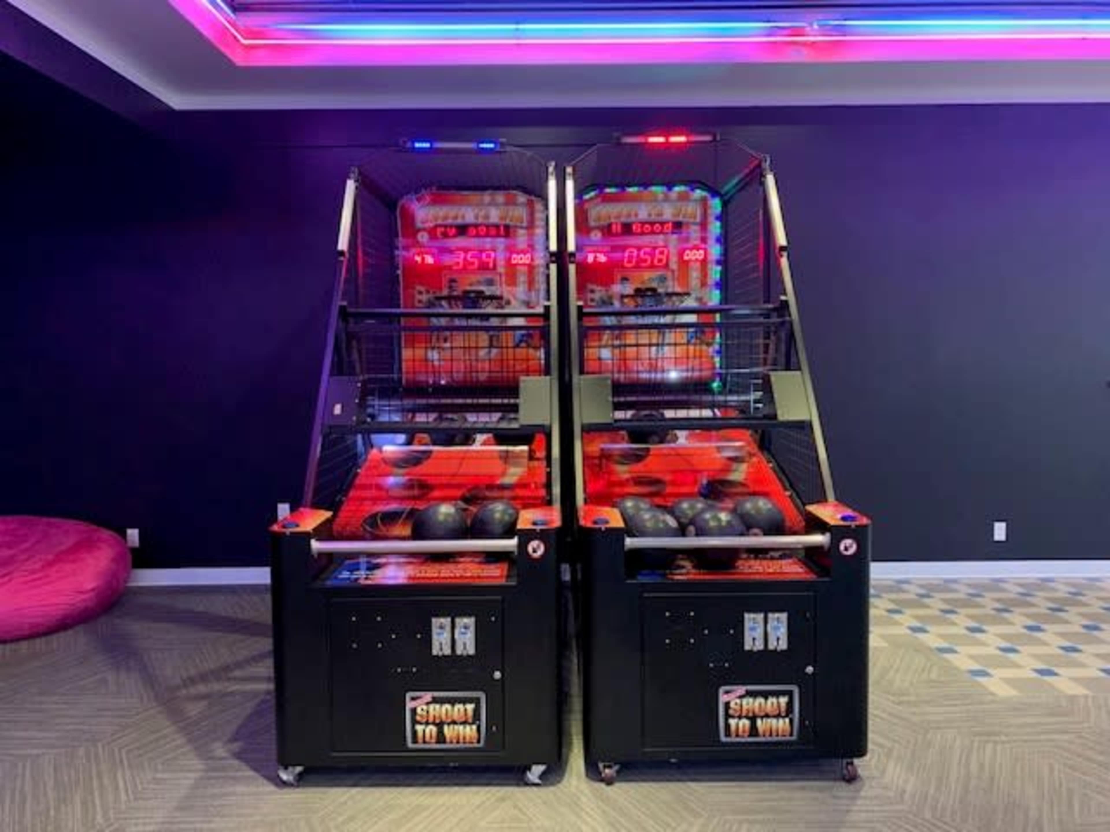 Two arcade basketball machines are set up against a dark wall, each displaying scores and illuminated with colorful lights.