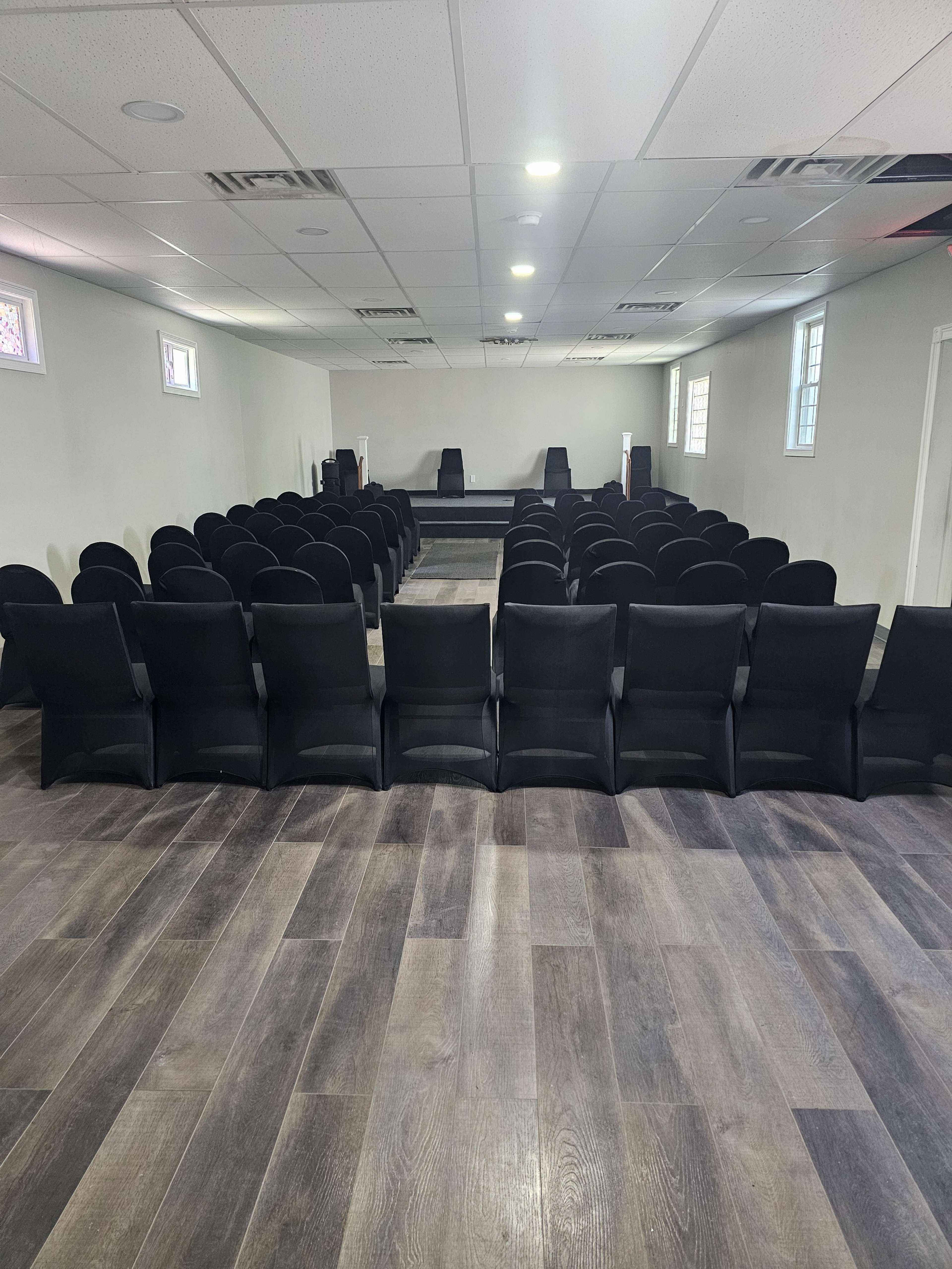 The image shows a room with rows of black chairs facing a small stage at the front.