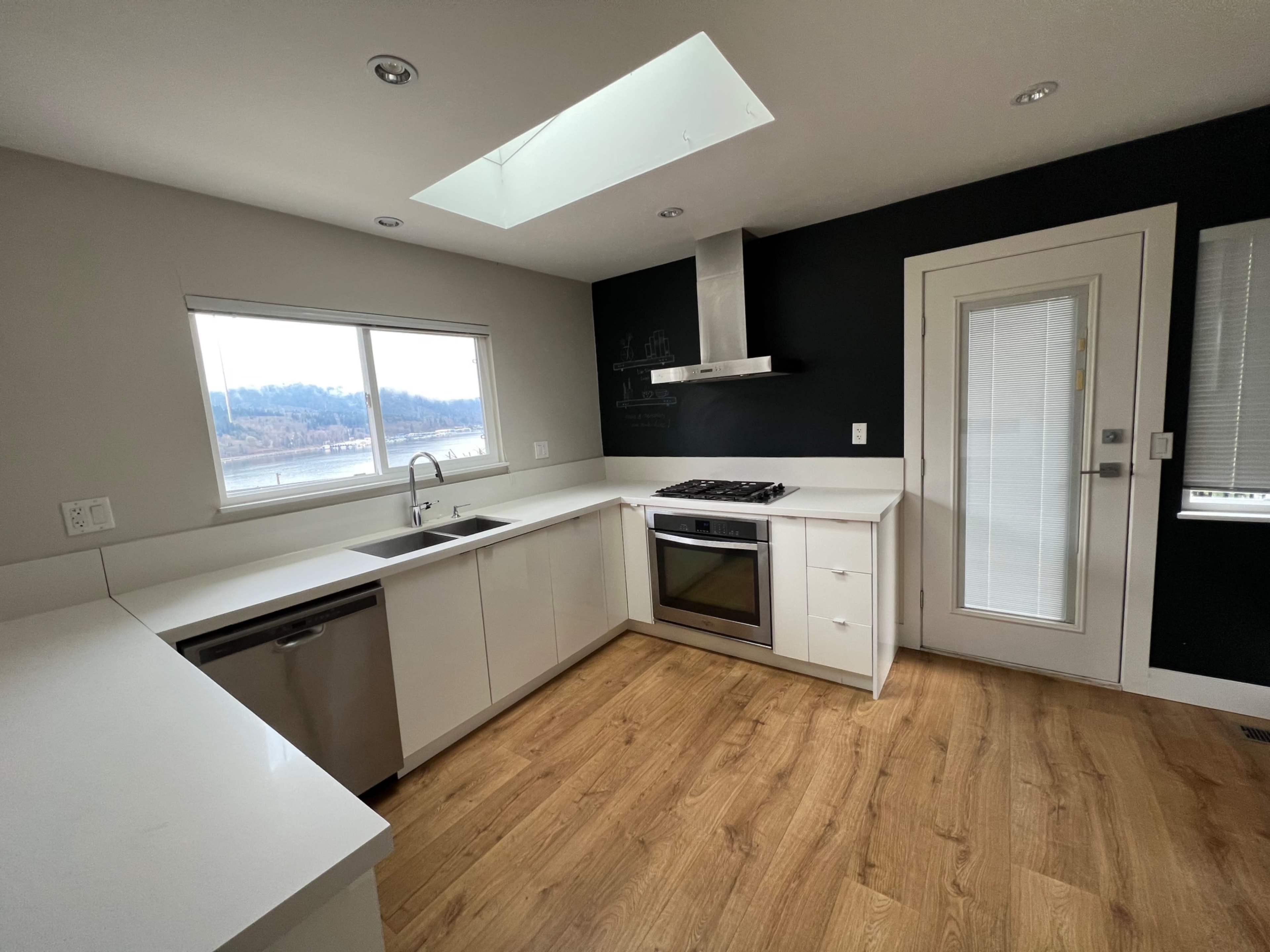A modern kitchen with white cabinetry, a stainless steel oven, a sink under a window, and a glass door leading outside.