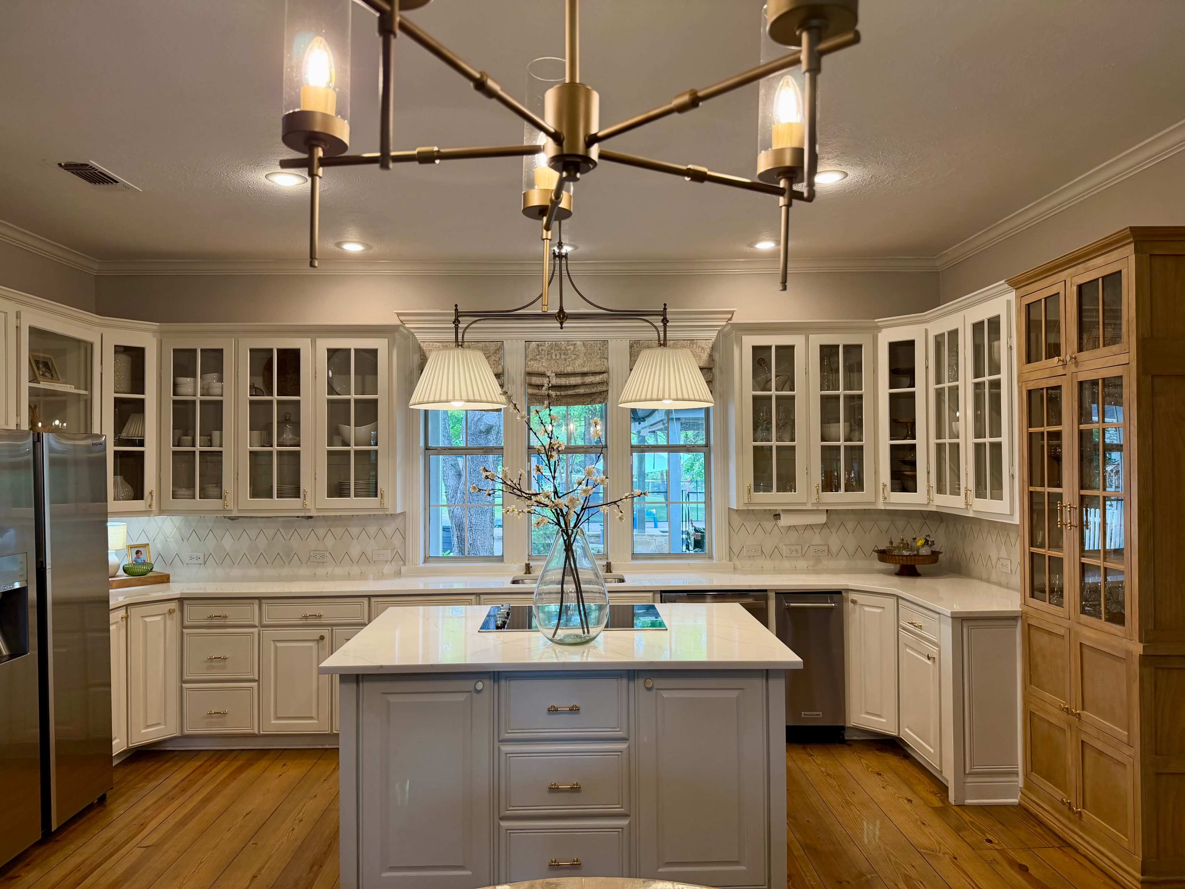 The image shows a spacious, modern kitchen featuring white cabinetry, a large island with a sink, and pendant lighting, with large windows allowing natural light to enter.